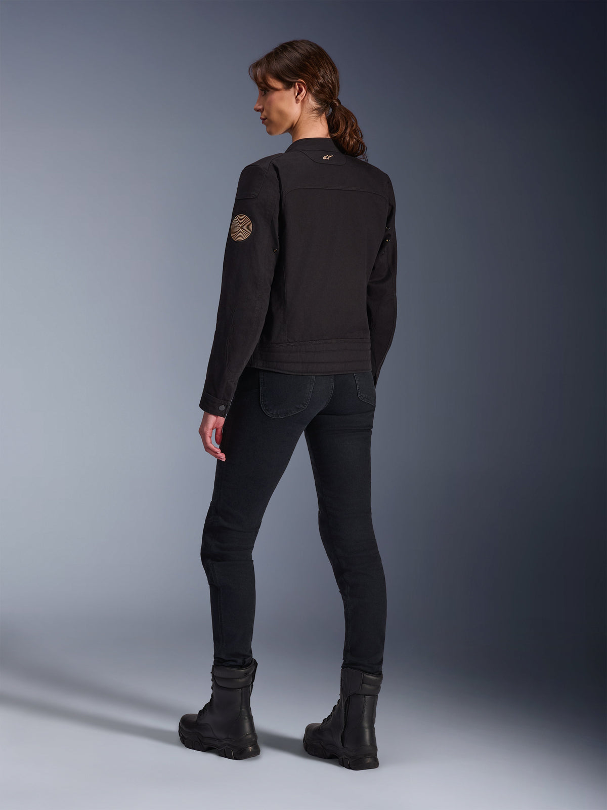 Modelo luciendo la chaqueta Stella Grace de Alpinestars en negro, vista posterior que destaca el parche espiral vintage Oscar en el brazo y el pequeño logo de estrella en el cuello, combinada con pantalones de moto negros y botas negras para conducción urbana