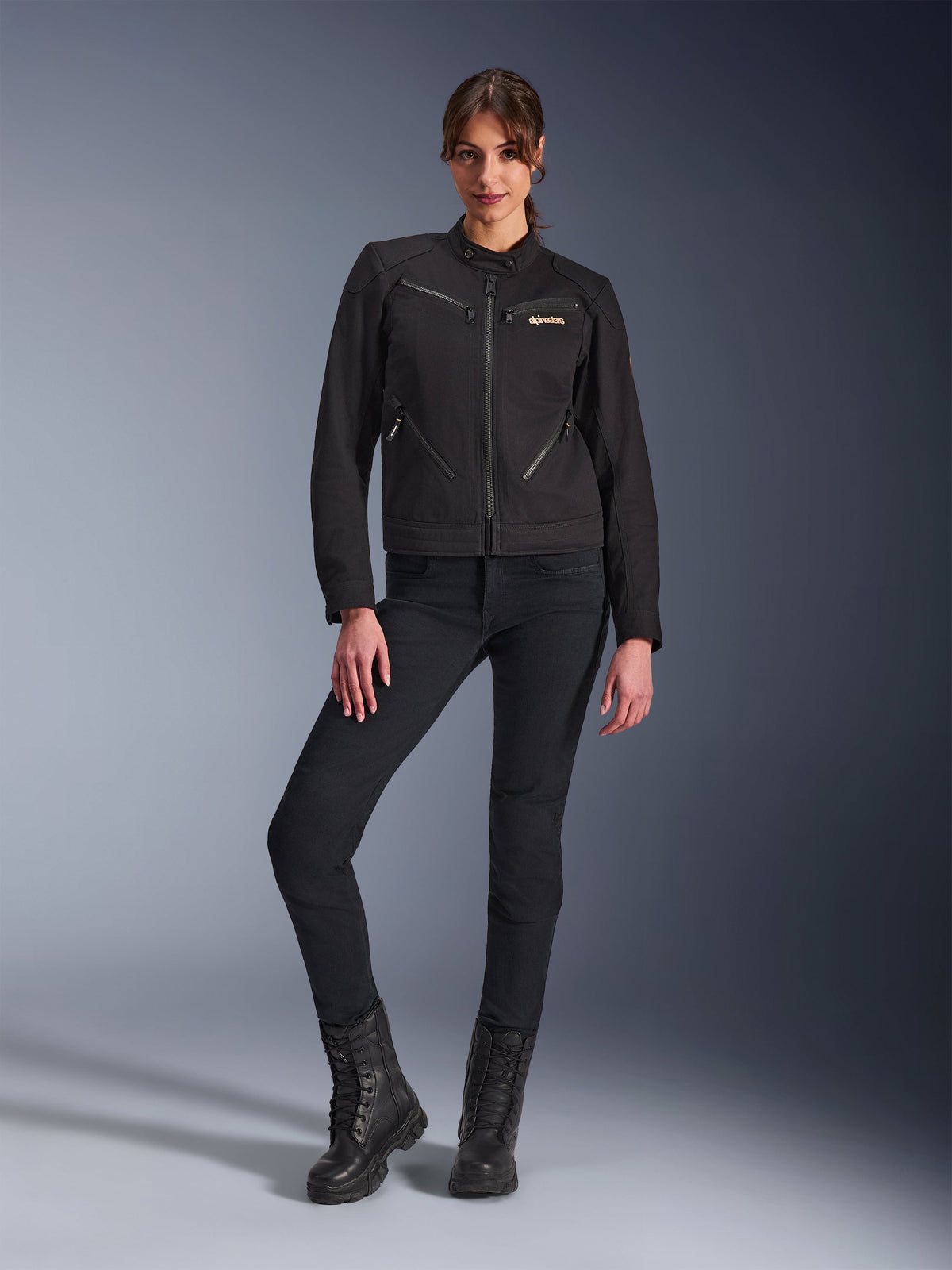 Mujer con la chaqueta Alpinestars Stella Grace en negro, chaqueta urbana de moto con estética de ropa de trabajo, con bolsillos frontales con cremallera y logotipo en el pecho, combinada con pantalones negros de corte ajustado y botas negras, equipamiento de motociclismo contemporáneo para mujer