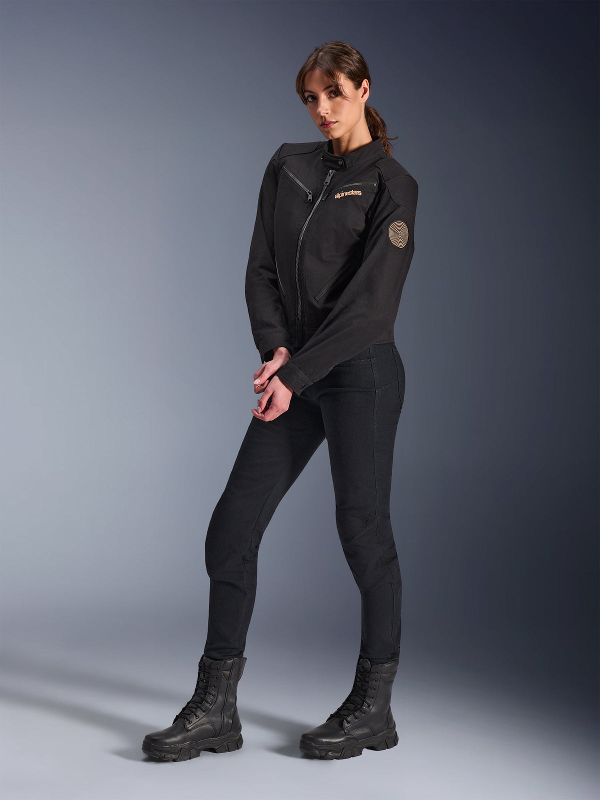 Modelo femenina con la chaqueta Stella Grace de Alpinestars para mujer, chaqueta urbana de moto negra, combinada con pantalones y botas de montar negros, diseño inspirado en la ropa de trabajo con logotipo bordado en bronce en el pecho y parche circular en espiral en la manga