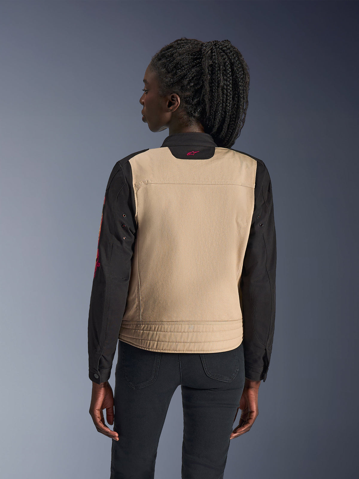 Chaqueta Alpinestars Stella Grace para mujer, chaqueta de moto urbana, Aluminum y Black, vista posterior que muestra un panel trasero color canela y mangas negras, diseño inspirado en la ropa de trabajo con un pequeño logo rosa en el cuello, combinada con pantalones oscuros