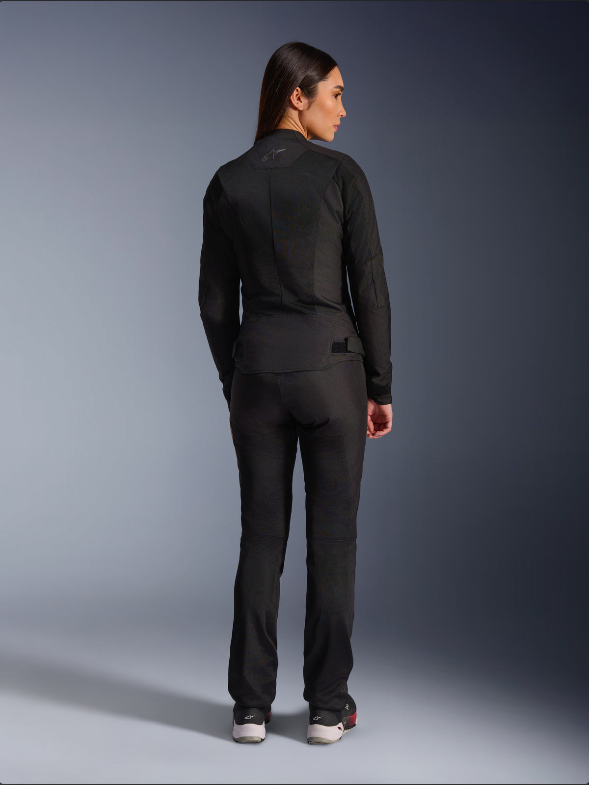 Chaqueta Alpinestars Stella C-1 Air, chaqueta de moto textil para mujer, negra, vista trasera mostrando la construcción de malla y un sutil logotipo en la parte superior de la espalda, combinada con pantalones de conducción negros a juego, estilo urbano contemporáneo