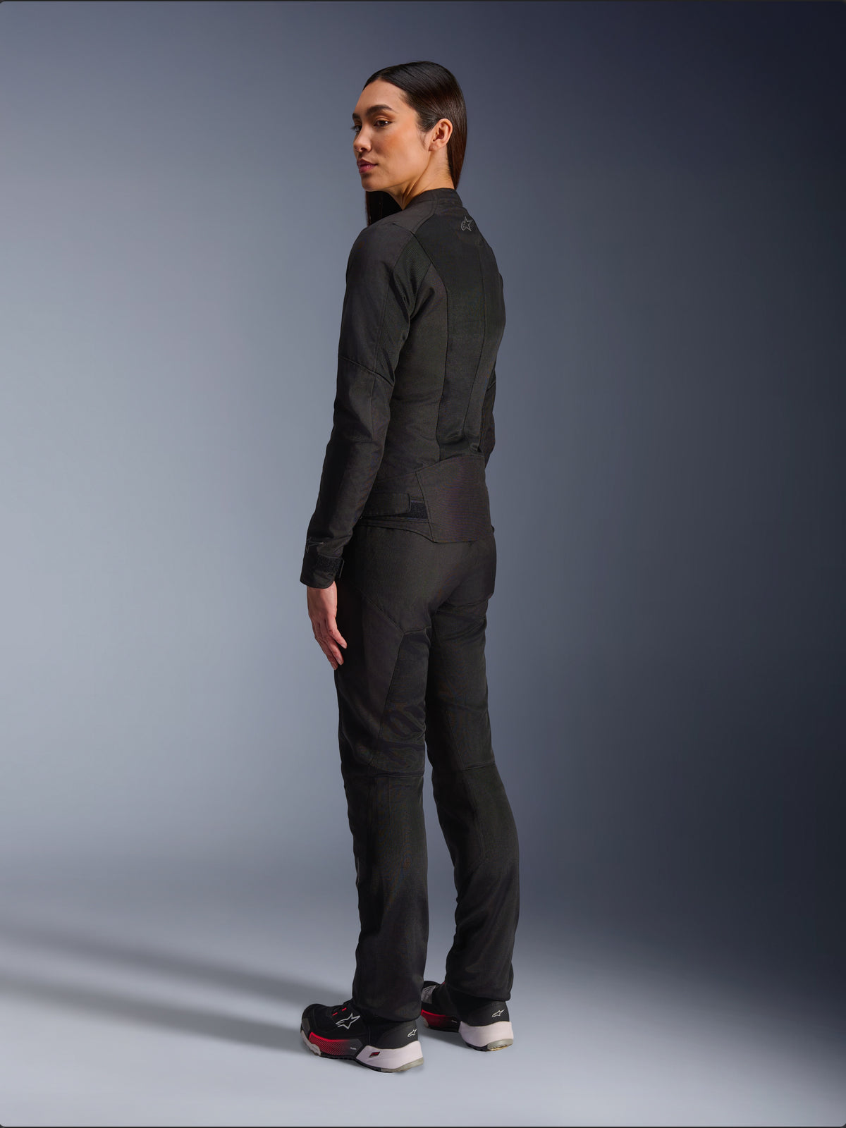 Mujer con la chaqueta Alpinestars Stella C-1 Air en color negro, chaqueta textil urbana para moto con paneles de malla, vista lateral trasera que muestra el ajuste estilizado y el logotipo en la parte superior de la espalda, combinada con pantalones negros a juego y calzado deportivo, equipamiento de moto contemporáneo.