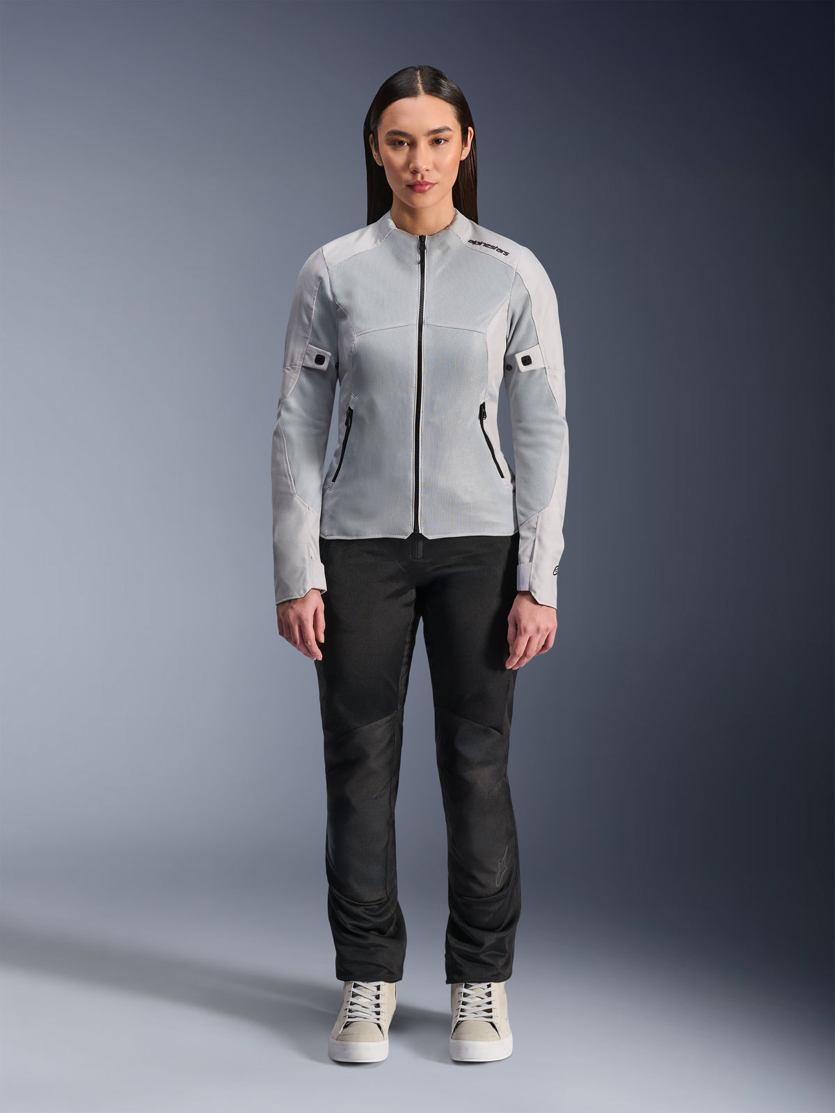 Mujer con chaqueta Alpinestars Stella C-1 Air en plateado, vista frontal, chaqueta de moto textil urbana con paneles de malla técnica, combinada con pantalones negros y zapatillas blancas, equipamiento de conducción contemporáneo