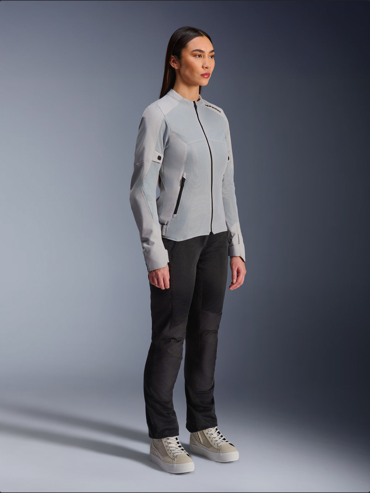Mujer vistiendo la chaqueta Alpinestars Stella C-1 Air en color plateado, chaqueta de moto de tejido ligero y malla, combinada con pantalones de conducción negros y zapatillas casuales, estética de conducción urbana limpia.