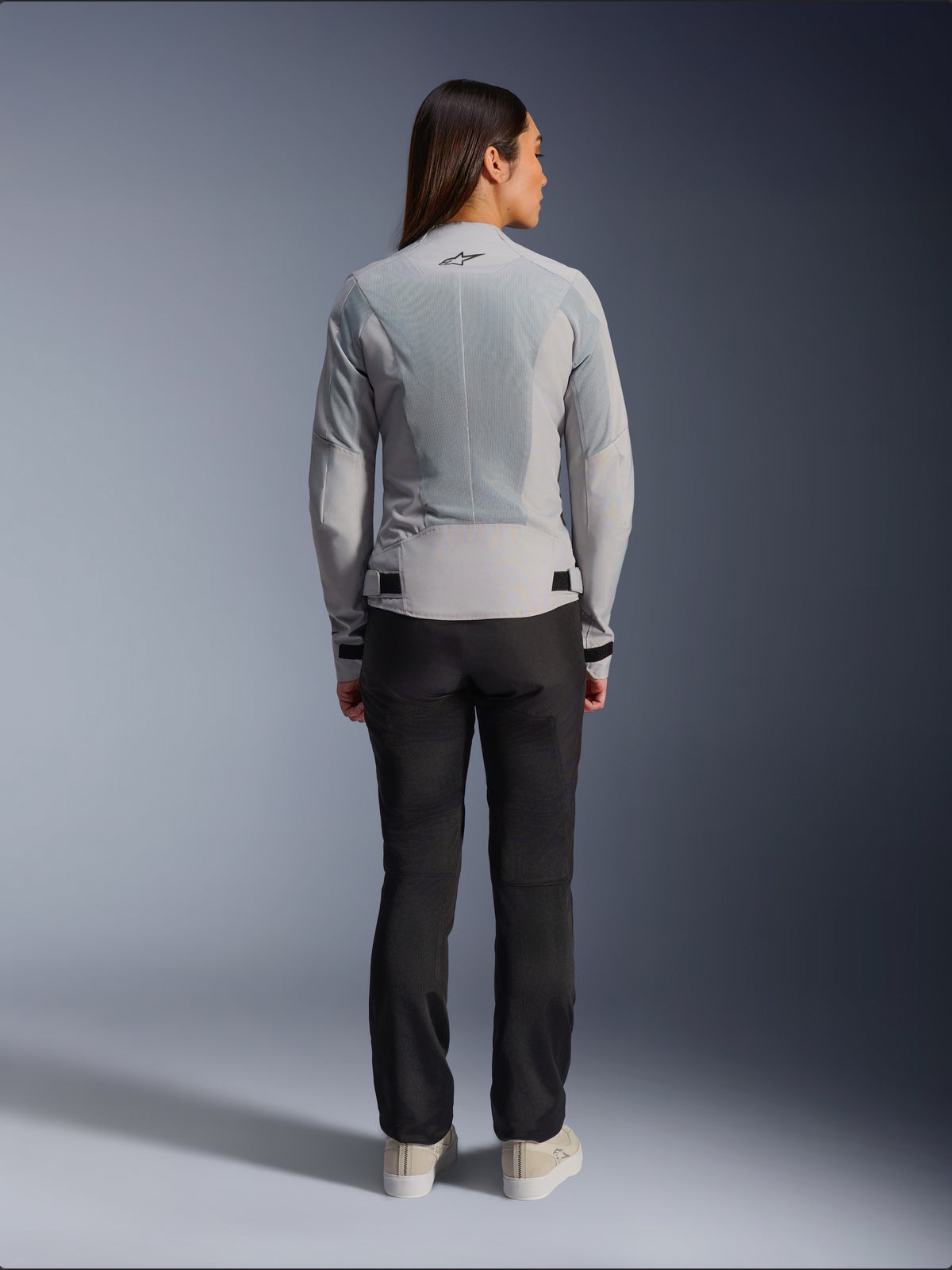 Mujer con la chaqueta Alpinestars Stella C-1 Air en color plateado, vista trasera, con grandes paneles de malla técnica para ventilación y un logotipo de estrella reflectante, combinada con pantalones de motociclista negros y zapatillas blancas, equipamiento de moto urbano contemporáneo