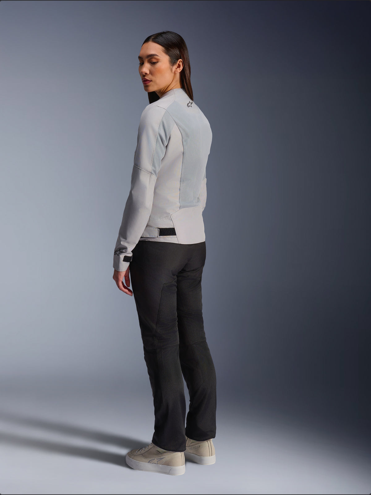 Mujer con la chaqueta Alpinestars Stella C-1 Air en plateado, vista trasera y lateral que muestra los paneles de malla técnica y el logotipo reflectante en el hombro, combinada con pantalones de moto negros y zapatillas beige, estética de conducción urbana contemporánea