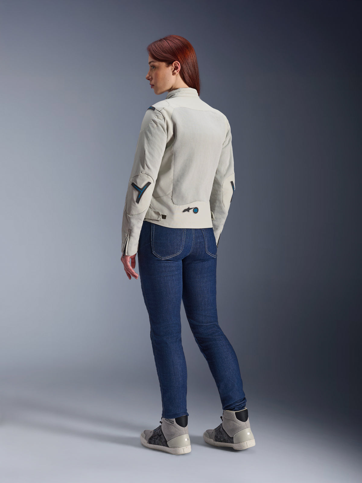 Mujer con la chaqueta Alpinestars Stella Circal-1 Air en gris metalizado y azul metilo, vista trasera que muestra los paneles de malla para el flujo de aire y detalles en azul neón, combinada con vaqueros de moto azul oscuro y zapatillas grises, toma de estilo de vida urbano para motociclismo.