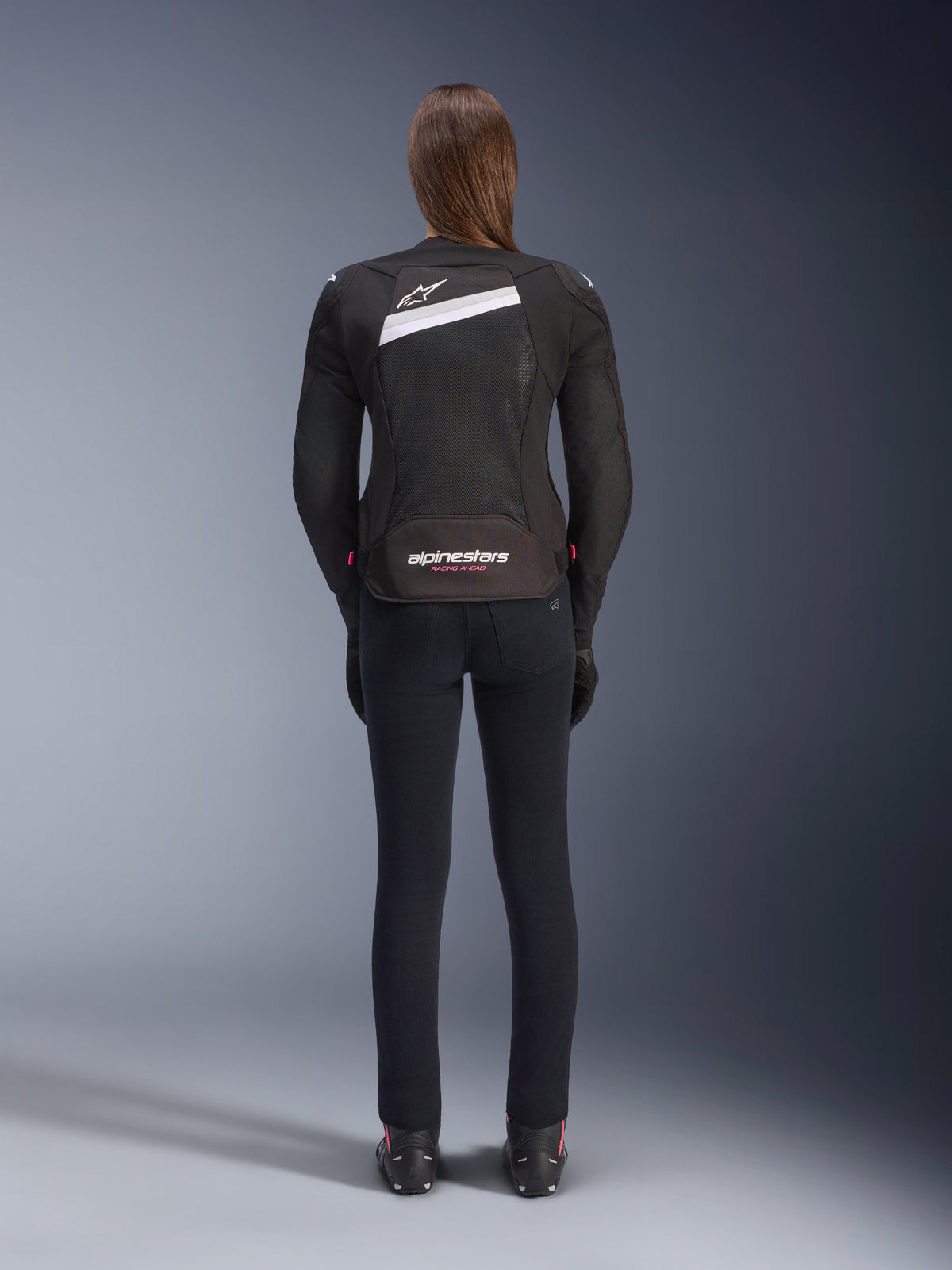 Alpinestars Stella T-GP Plus R V4 Airflow Jacket, Black and White, rear view showing extensive mesh ventilation panels and Alpinestars branding, worn by a female rider with slim-fit black motorcycle pants and boots, sport-fit textile gear for women