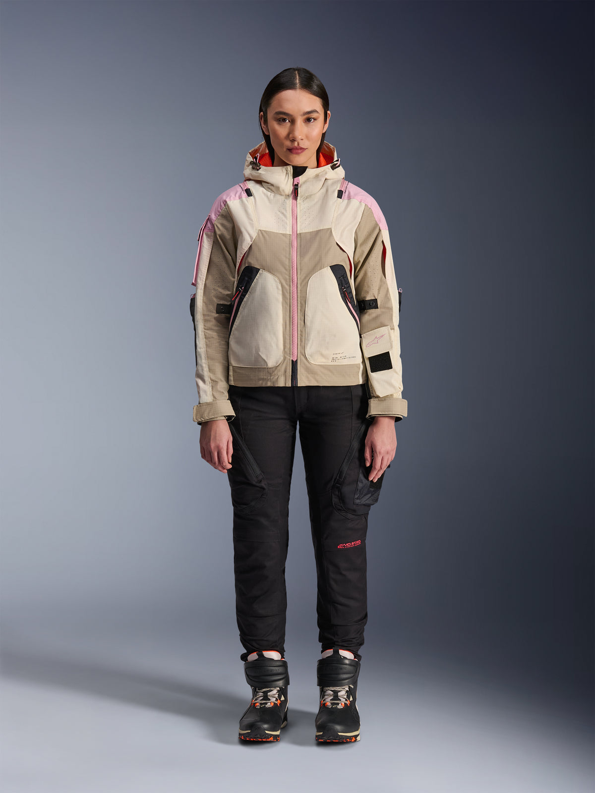 Modelo femenina con la chaqueta Alpinestars Molly Air en color Sand Pink Almond, combinada con pantalones técnicos de motociclismo y botas negros, vista frontal de cuerpo completo, chaqueta de moto urbana con diseño de inspiración utilitaria y detalles en rosa.