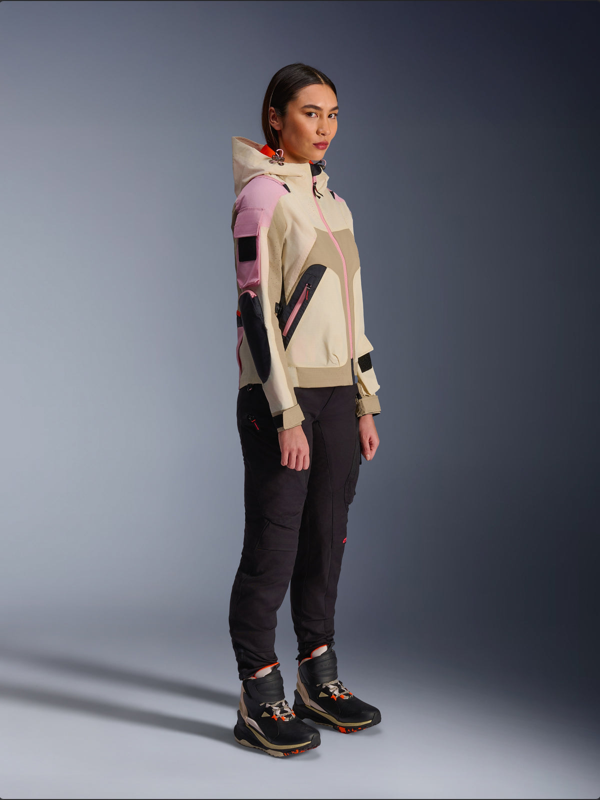 Modelo femenina luciendo la chaqueta Alpinestars Molly Air en Sand Pink Almond, combinada con pantalones técnicos oscuros y botas de motociclismo de caña alta, una chaqueta de moto urbana con capucha y paneles multicolores con diseño contemporáneo inspirado en el estilo streetwear