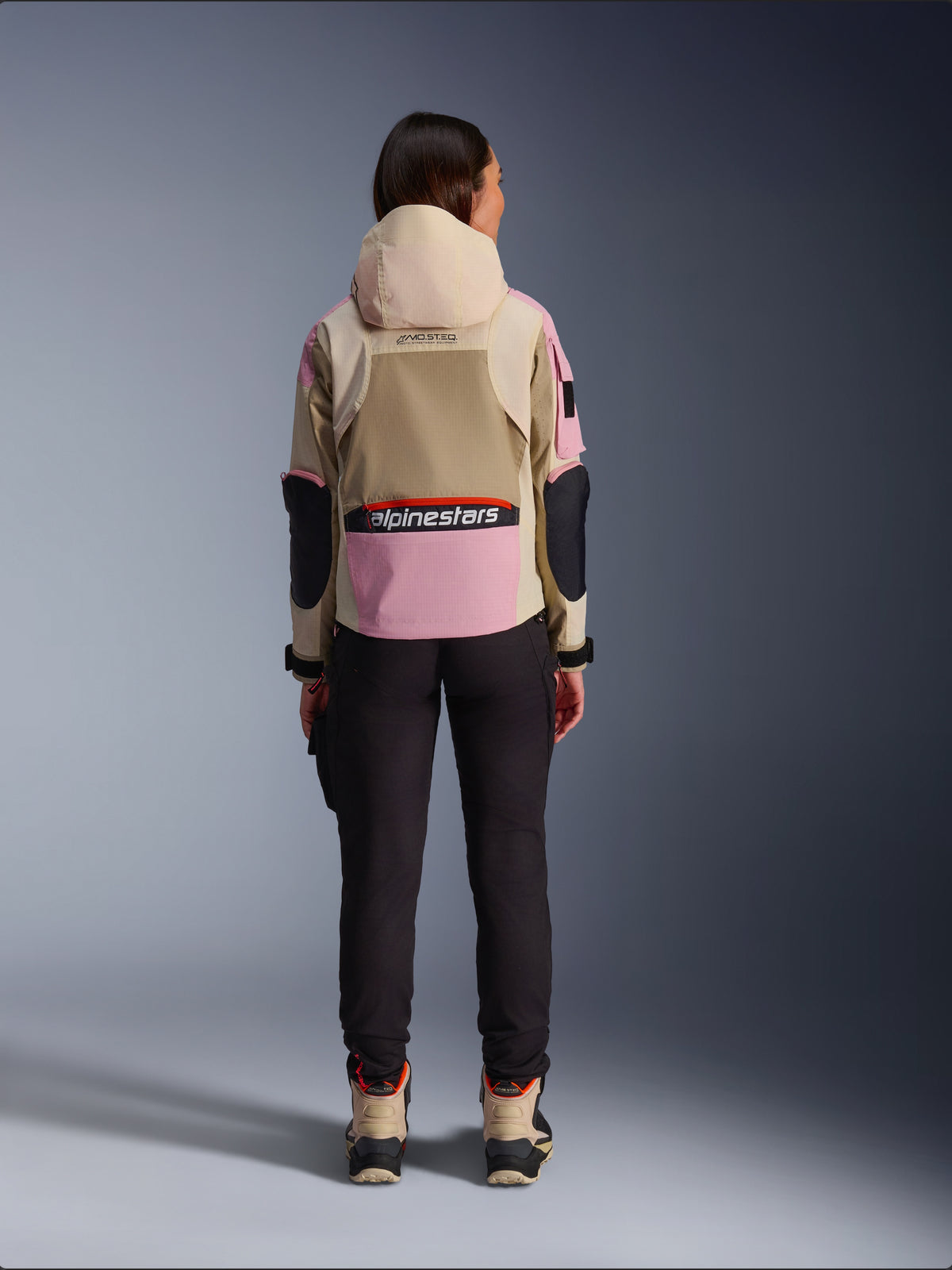Female rider viewed from behind wearing the Alpinestars Molly Air Jacket in Sand Pink Almond, featuring a tan hood and pink panels with black accents, paired with black riding pants and tan motorcycle boots, urban contemporary motorcycle apparel