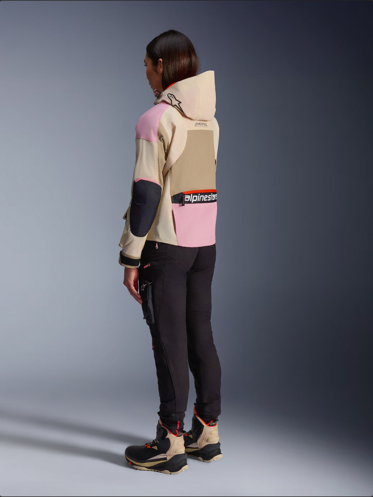 Motociclista con la chaqueta Alpinestars Molly Air en Sand Pink Almond, con un diseño multitonal en beige y rosa, vista lateral trasera con pantalones de moto negros y botas técnicas a juego, estilo de motociclismo urbano con capucha.