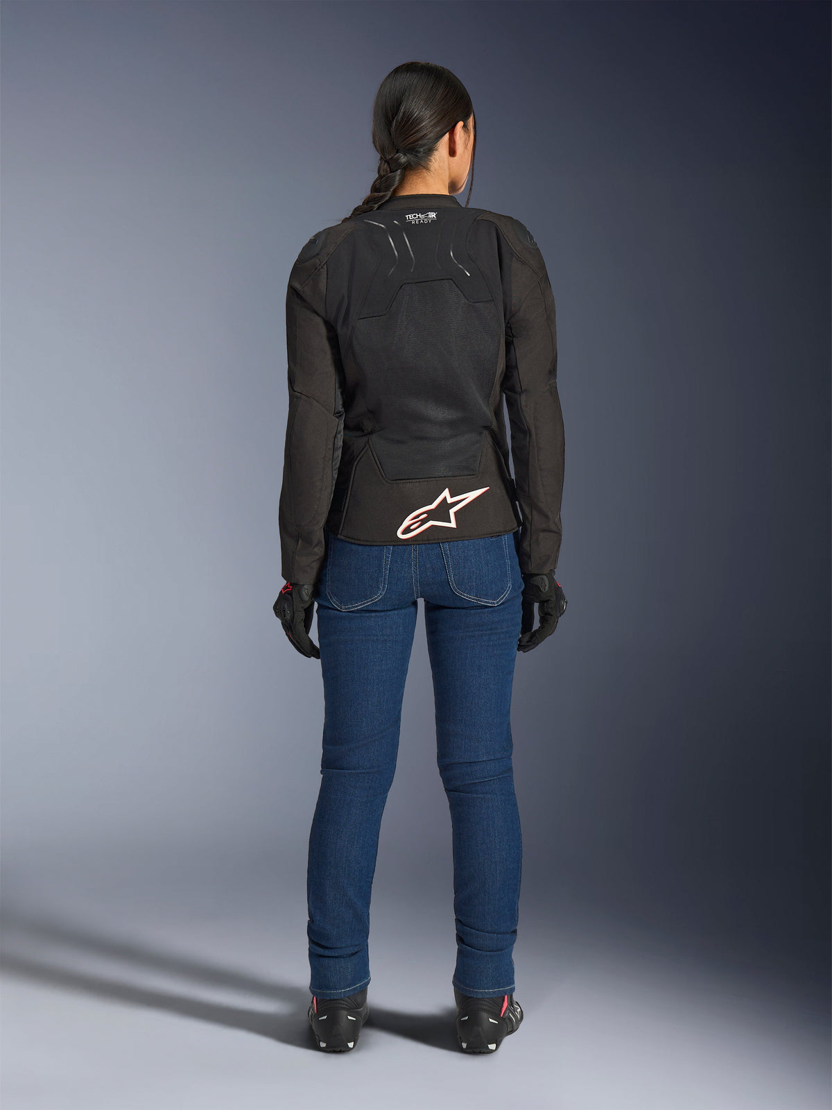 Female rider from the back wearing the Alpinestars Stella SMX Air Jacket in Black, paired with blue denim riding pants and black gloves, featuring a ventilated textile design, white star logo on the lower back, and Tech-Air Ready branding, designed for warm-weather sport riding