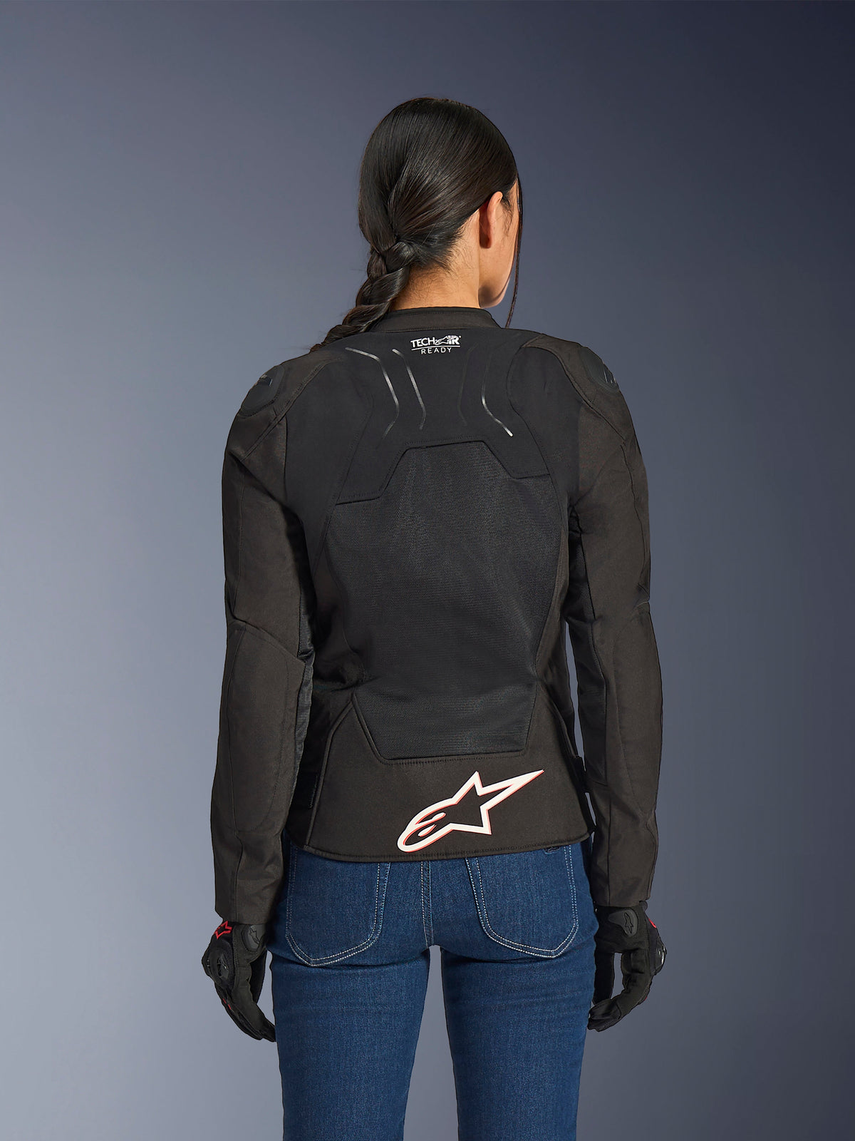 Alpinestars Stella SMX Air Jacket, back view on female model, black textile with high-flow mesh panels, 'TECH-AIR READY' branding on the upper back, large white Alpinestars logo with red outline on the lower back, paired with blue denim jeans and black riding gloves