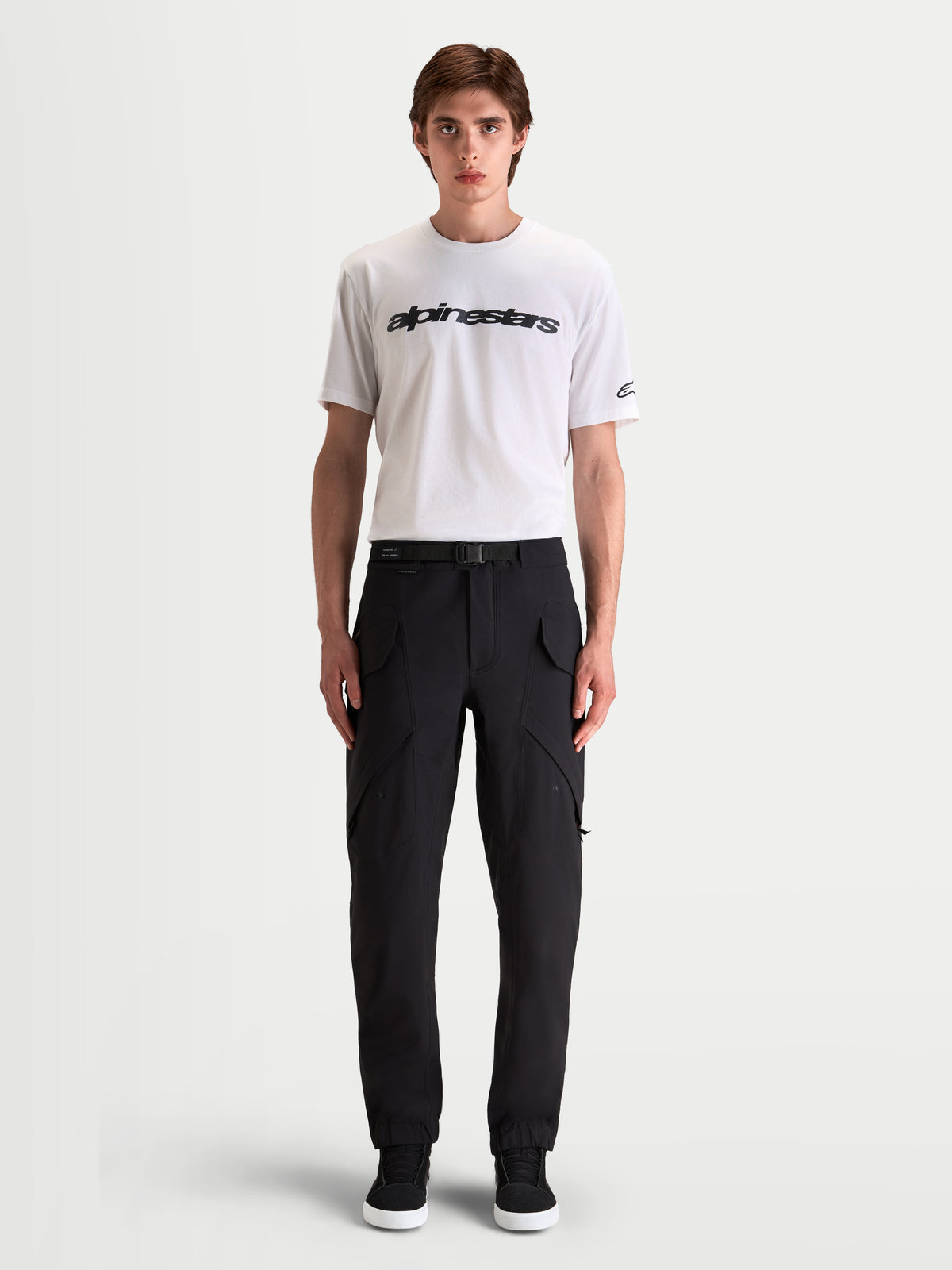 Alpinestars Juggernaut V2 Drystar Pantalones, pantalones de moto urbanos para hombre, negros, vista frontal en modelo con camiseta blanca y botas casuales, con bolsillos cargo y cintura con cinturón ajustable