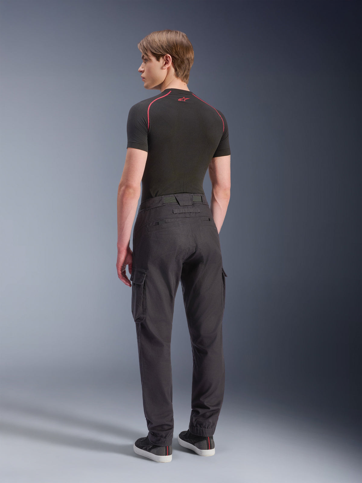 Alpinestars Flex-AST Cargo Pants - Short, pantalones cargo de moto, negros, vista trasera de modelo con pantalones cargo de ajuste regular tapered, bolsillos laterales y dobladillos elásticos, combinados con una capa base negra y calzado casual