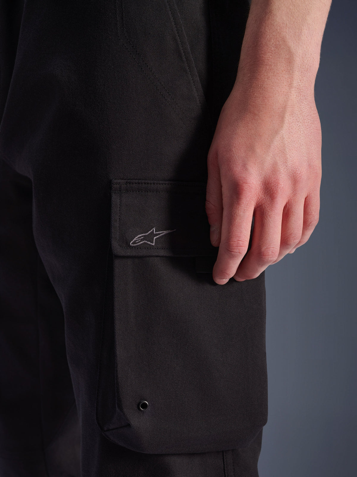 Alpinestars Flex-AST Cargo Pants, pantalones de moto urbanos, negros, primer plano del bolsillo cargo lateral con logo Astar bordado, ojal metálico y textura de sarga elástica duradera