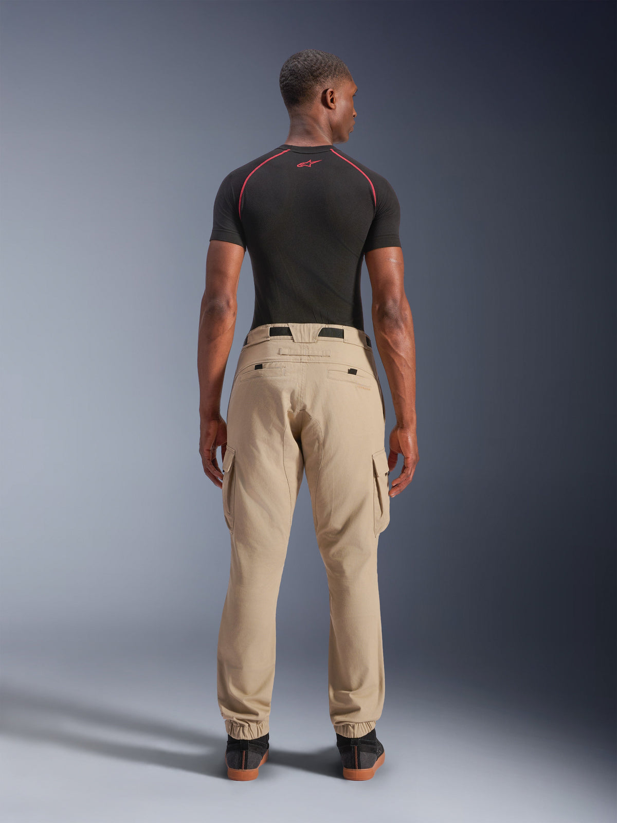 Pantalones Cargo Alpinestars Flex-AST - Cortos, beige claro, vista trasera en un modelo, ajuste regular tapered con dobladillos elásticos y bolsillos cargo, combinados con una capa base técnica negra con detalles rojos, ropa de moto urbana moderna