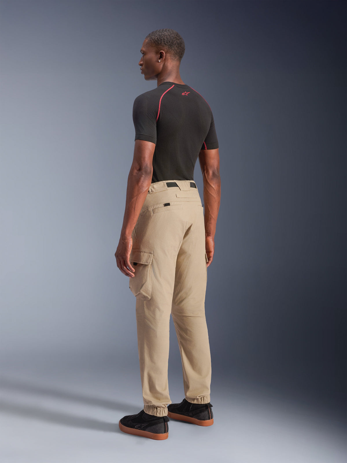 Alpinestars Flex-AST Cargo Pants, pantalones urbanos para moto, Beige claro, vistos desde un ángulo lateral trasero en un modelo, con bolsillos cargo funcionales y puños elásticos, combinados con una camiseta negra de manga corta y zapatillas negras.