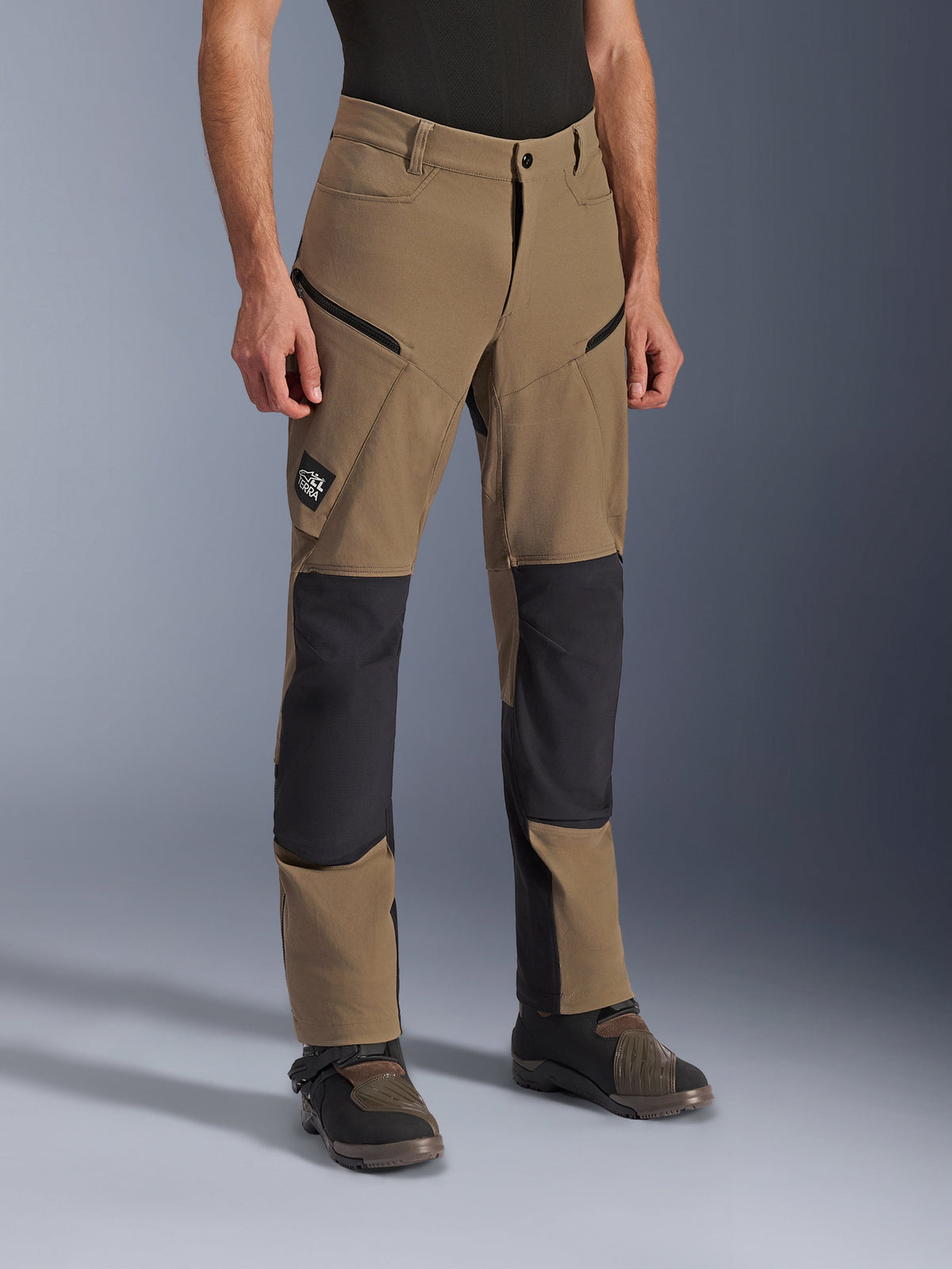 Pantalones Alpinestars Flex-AST Explorer, pantalones de motociclismo de aventura en Walnut Black, con textil canela y paneles reforzados negros en rodillas y asiento, bolsillos cargo con cremallera y parche con el logo All Terra, mostrados con botas técnicas de conducción.