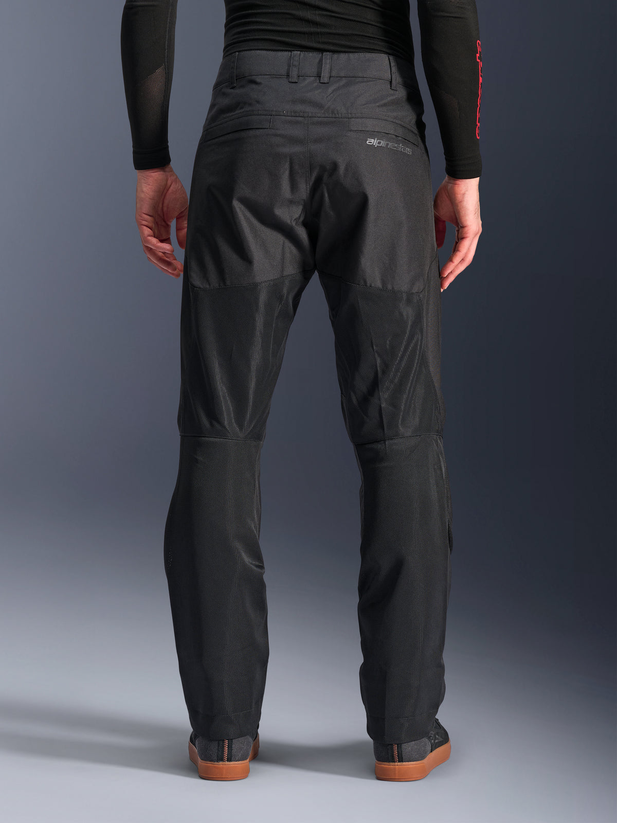 Pantalones Alpinestars C-1 Air, vista trasera, negros, pantalones urbanos de moto fabricados en tejido textil con amplios paneles de malla técnica en la parte posterior de las piernas para mayor ventilación, diseño limpio con marca sutil, combinados con una capa base negra y calzado casual para conducir.