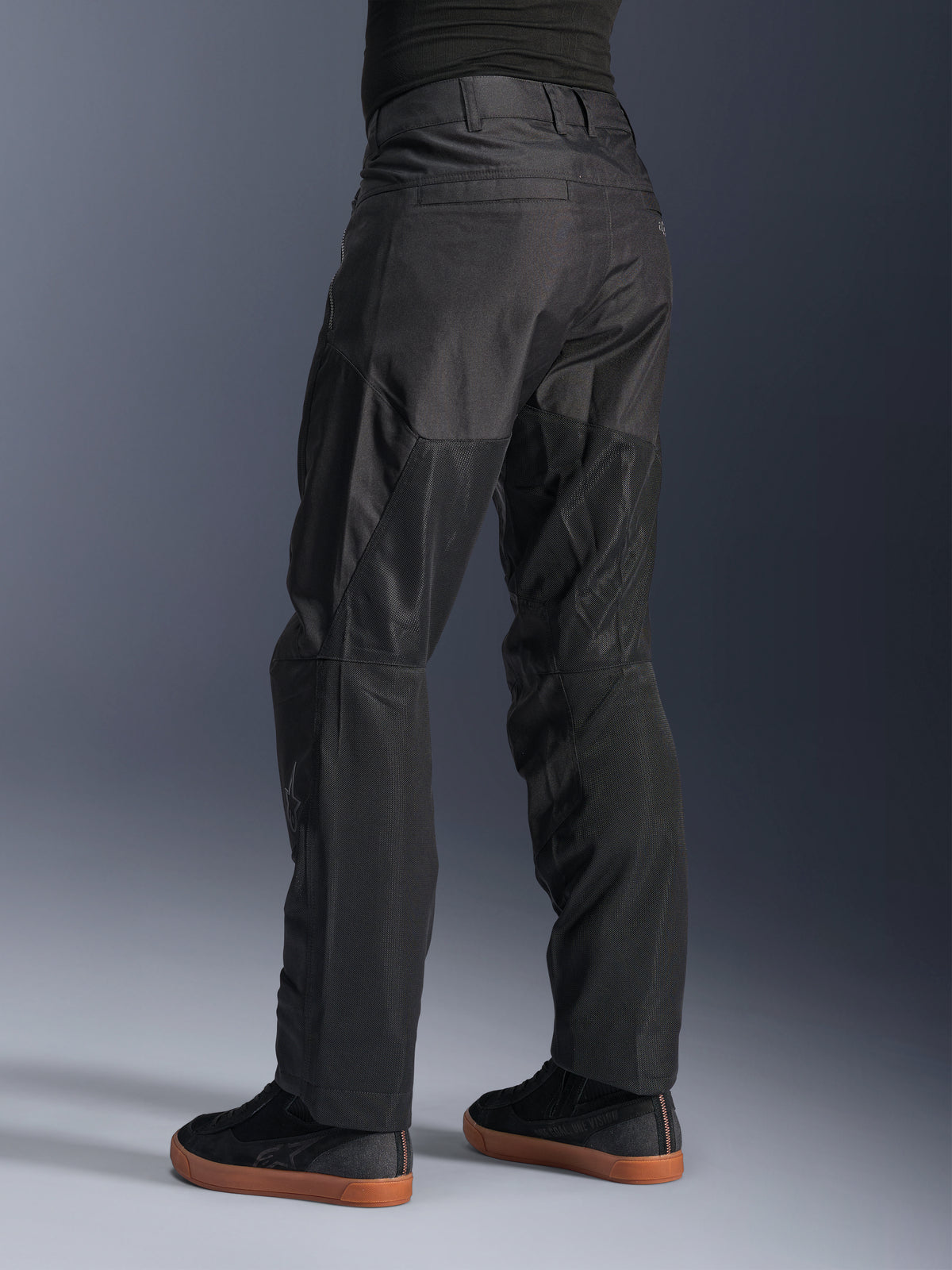 Alpinestars C-1 Air Pants, pantalones urbanos de moto, color negro, vista trasera que muestra grandes paneles de malla técnica en la parte posterior de las piernas para ventilación, estilo limpio para conducción urbana, combinados con zapatillas casuales negras.