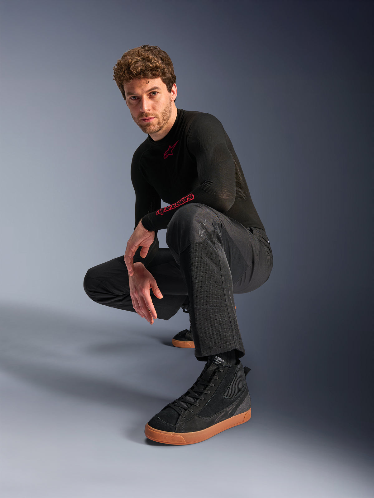 Pantalones Alpinestars C-1 Air, pantalones de moto urbanos, negros, modelo agachado en un estudio, combinados con una camiseta negra de manga larga y zapatillas negras con suela color canela, con paneles de malla transpirable y un estilo urbano contemporáneo