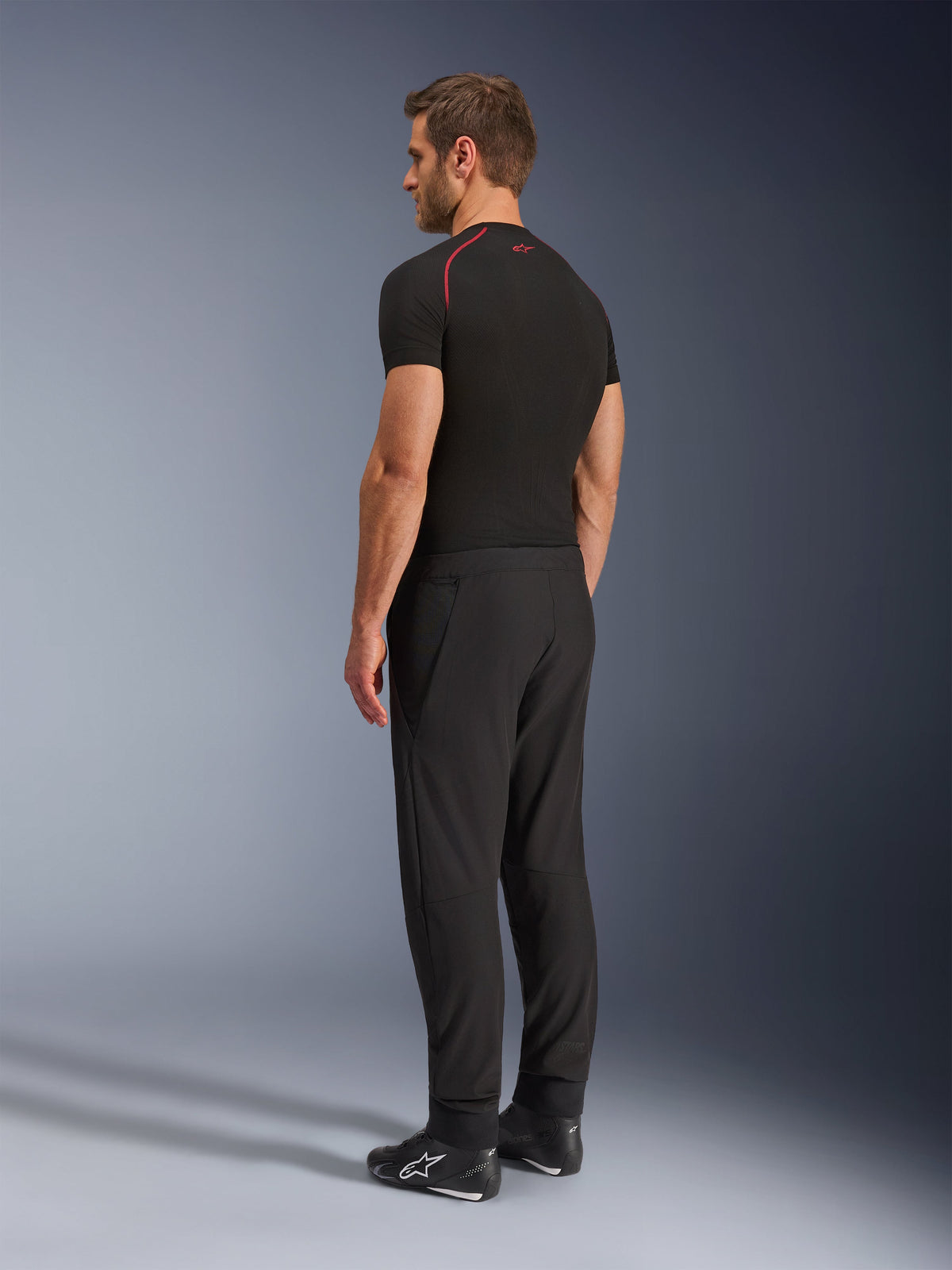Pantalones Alpinestars Aeroshell Airflow, pantalones de moto negros, tejido softshell ligero, usados con una camiseta técnica negra de manga corta y botas de conducción negras, vista de perfil lateral trasera que muestra el corte ajustado y la construcción ergonómica.