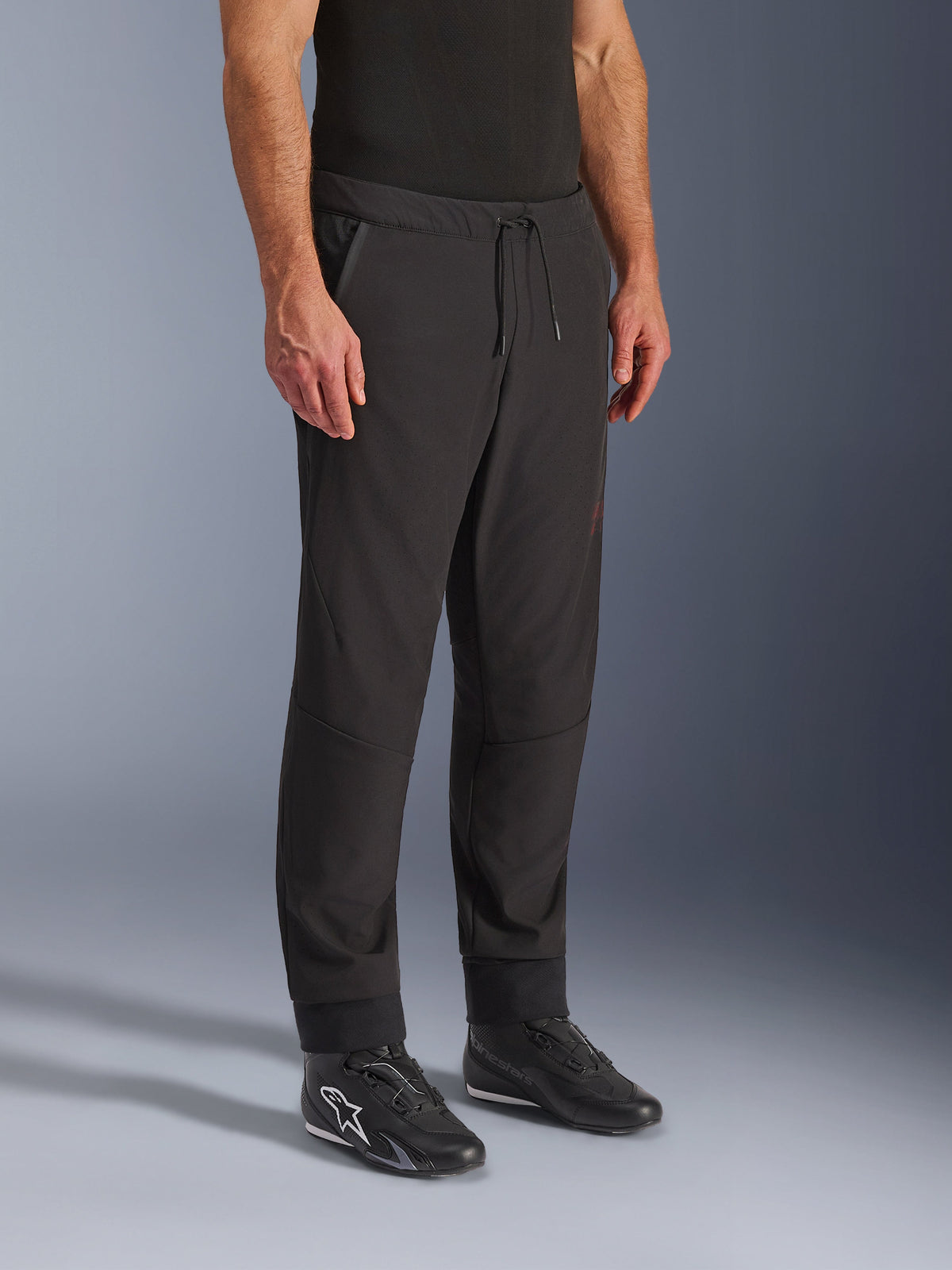 Pantalones Alpinestars Aeroshell Airflow, pantalones de motociclismo negros, usados por un modelo con calzado de conducción negro, con cintura con cordón ajustable, puños en los tobillos y tejido softshell perforado con láser para una máxima ventilación