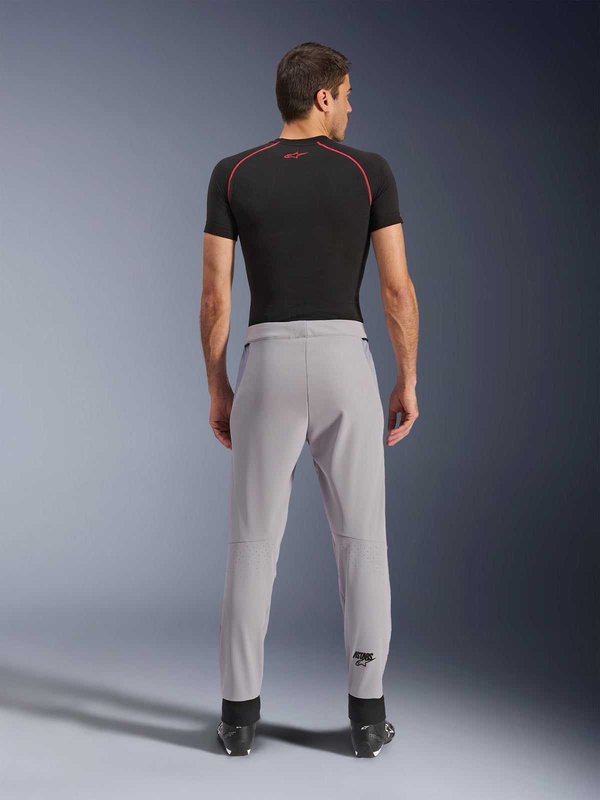 Pantalones Alpinestars Aeroshell Airflow, Gris Medio, vista trasera que muestra las zonas perforadas con láser en las pantorrillas, construcción de softshell elástico ligero, usados con una camiseta negra de manga corta y botas de moto, diseño de ajuste deportivo para climas cálidos