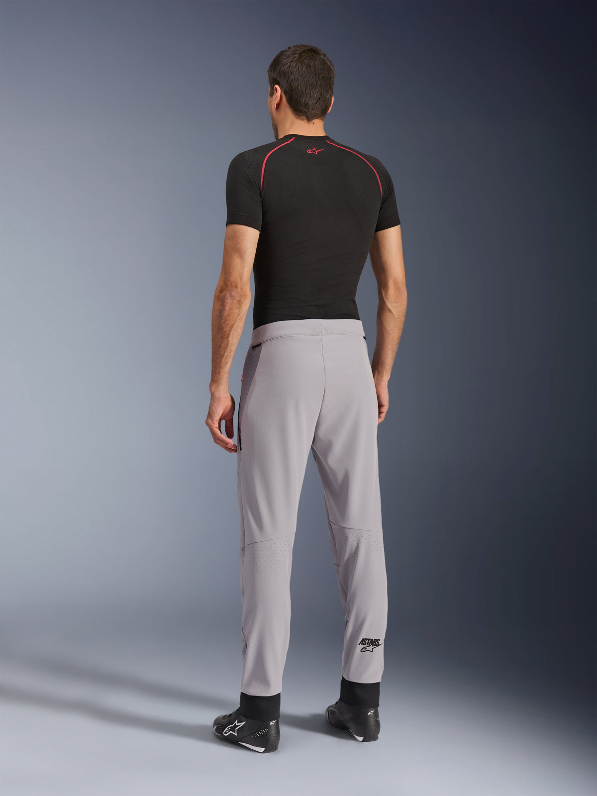 Vista posterior de un modelo con los pantalones Alpinestars Aeroshell Airflow en color Mid Gray, combinados con una camiseta técnica negra y calzado negro, destacando las zonas perforadas con láser en las pantorrillas y el ajuste deportivo atlético.
