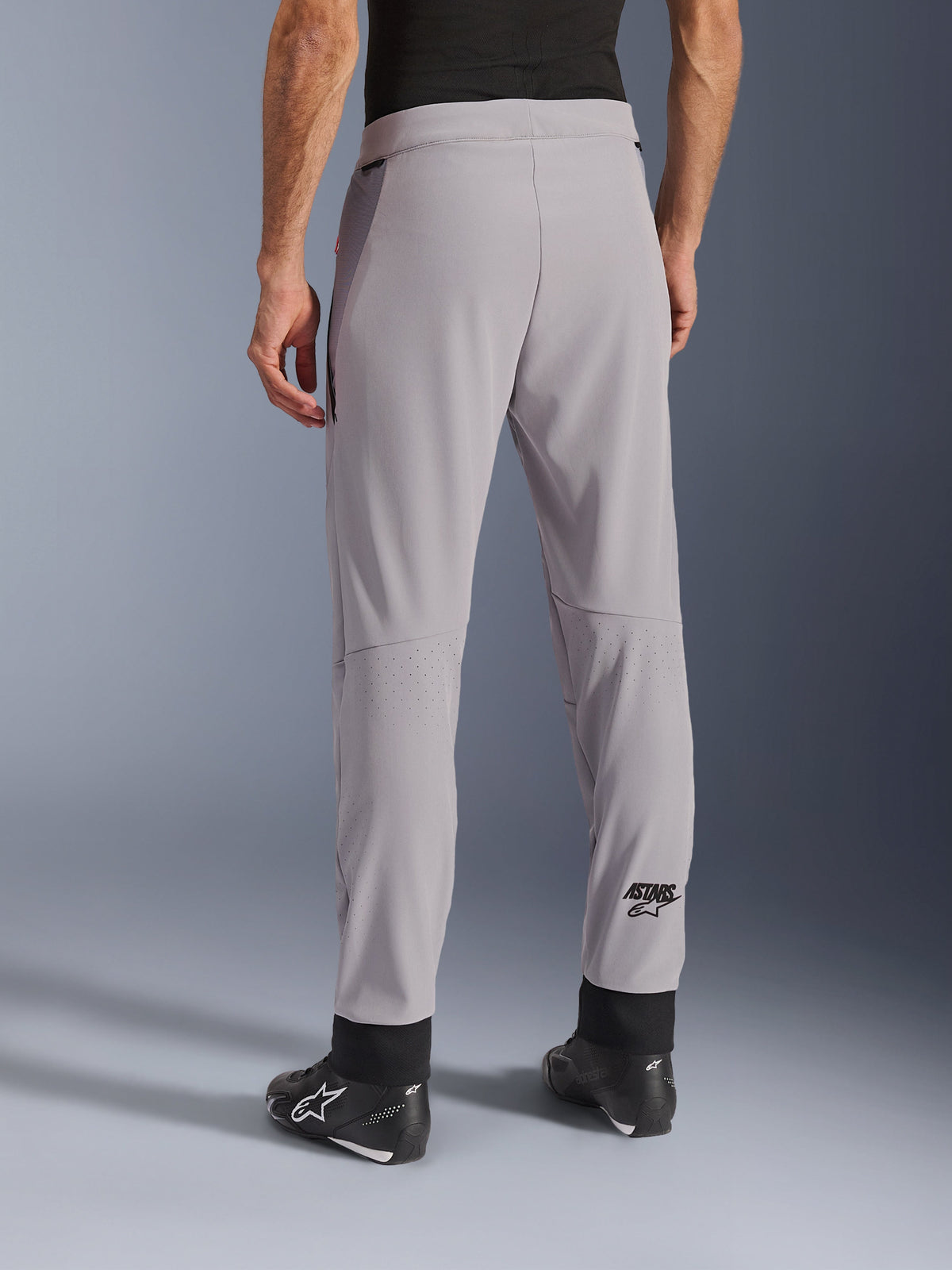 Pantalones Alpinestars Aeroshell Airflow, Gris Medio, vista trasera que muestra zonas perforadas con láser en las pantorrillas, logotipo ASTARS en la parte inferior de la pierna, puños de tobillo ajustables negros, combinados con botas de moto negras, pantalones textiles ligeros para conducción deportiva