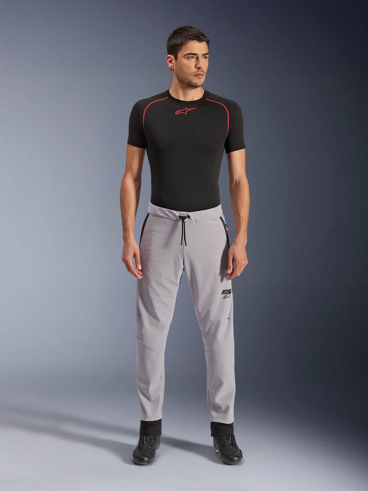 Pantalones Alpinestars Aeroshell Airflow, Gris Medio, pantalones textiles ligeros con zonas de ventilación perforadas con láser y cintura con cordón, usados por un modelo con una camiseta de compresión negra y botas de moto negras, diseño de conducción deportiva limpio y moderno.