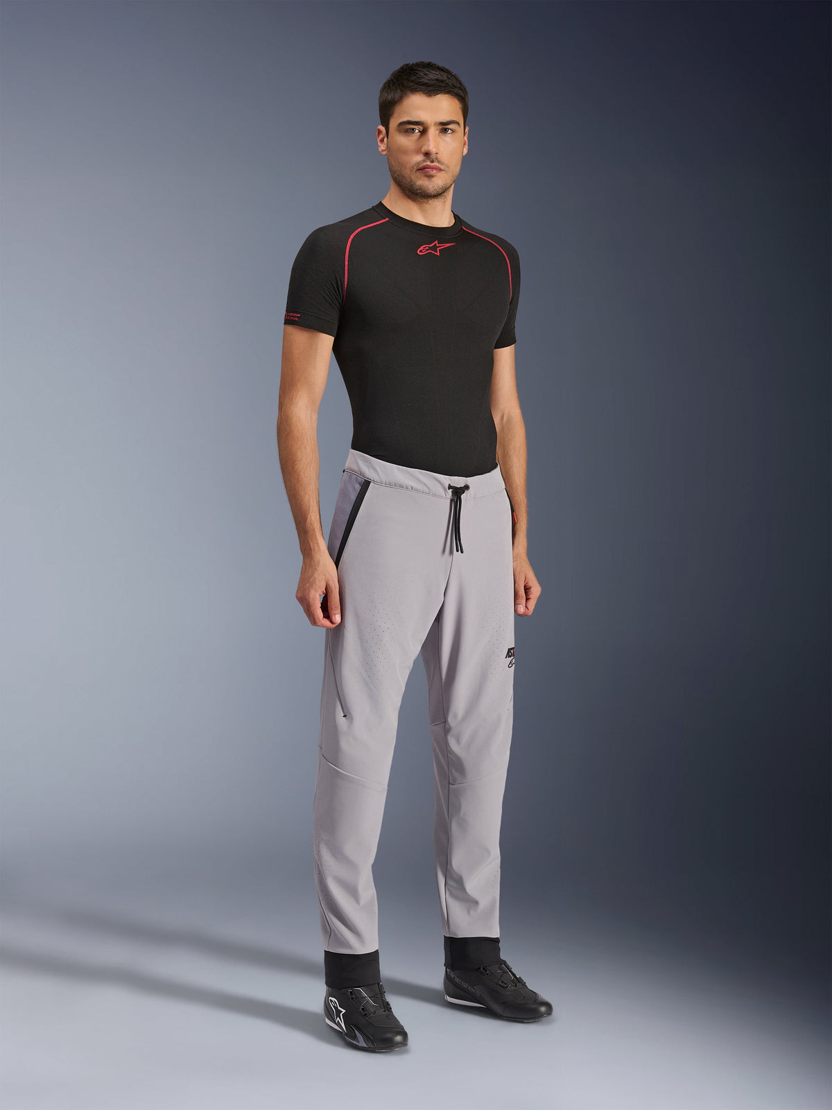 Pantalones Alpinestars Aeroshell Airflow, pantalones de moto, gris medio, usados por un modelo con una camiseta técnica negra y calzado de conducción negro, con zonas de ventilación perforadas con láser, cintura elástica con cordón y un ajuste deportivo estilizado.