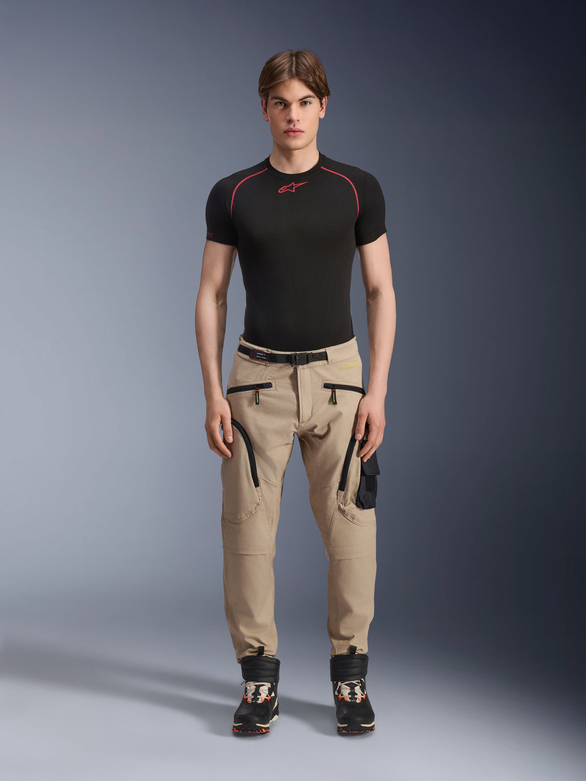 Modelo masculino vistiendo los pantalones Alpinestars Acteon en color Sand, combinados con una capa base negra de manga corta y botas técnicas negras, con bolsillos cargo con cremalleras negras y un ajuste entallado, equipamiento contemporáneo de moto urbana.