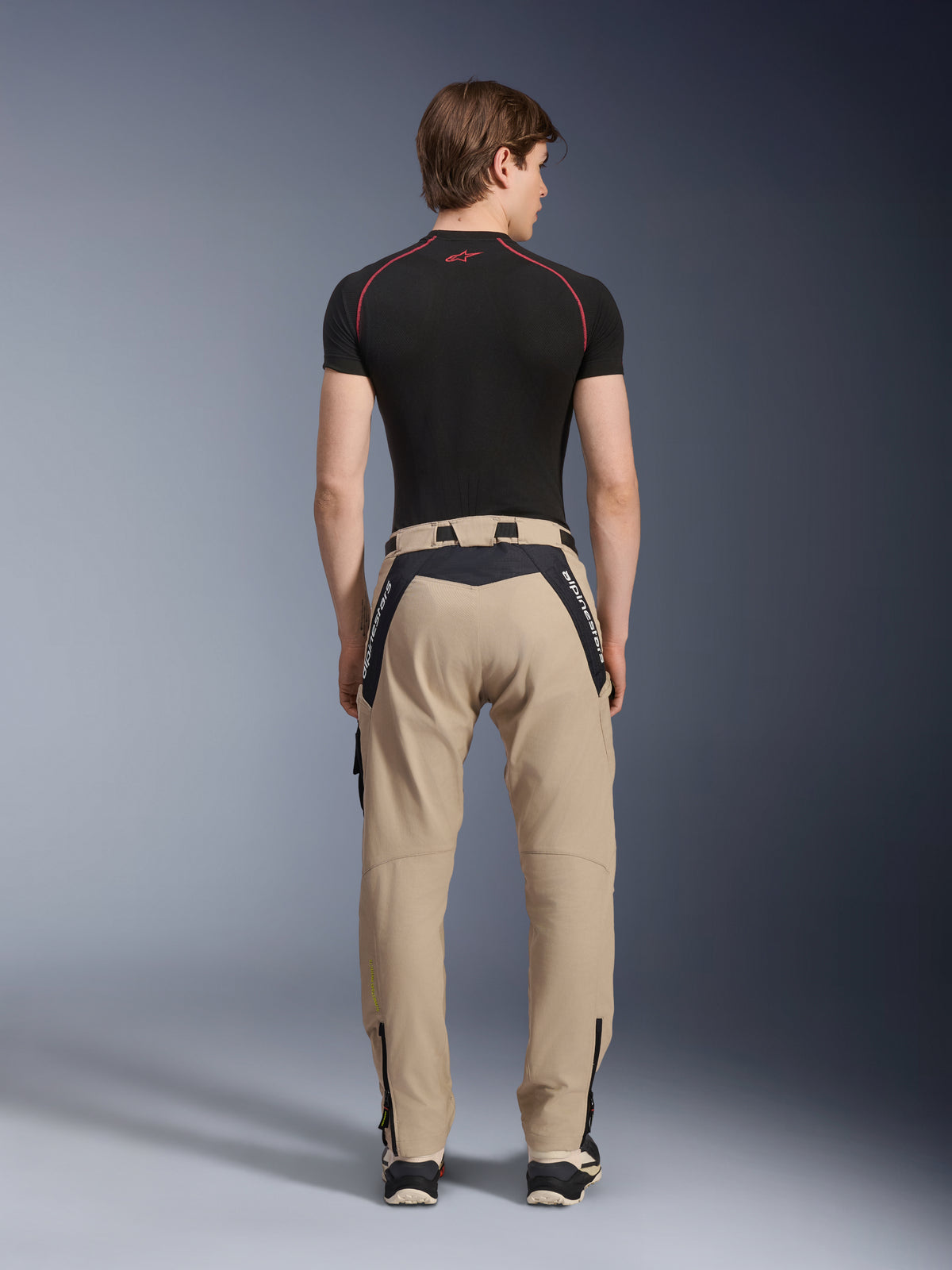 Pantalones Alpinestars Acteon en color Arena, modernos pantalones de moto urbanos vistos desde atrás, con paneles negros y la marca Alpinestars en blanco, corte ajustado con cremalleras en los bajos, combinados con una prenda técnica de manga corta negra y zapatillas.
