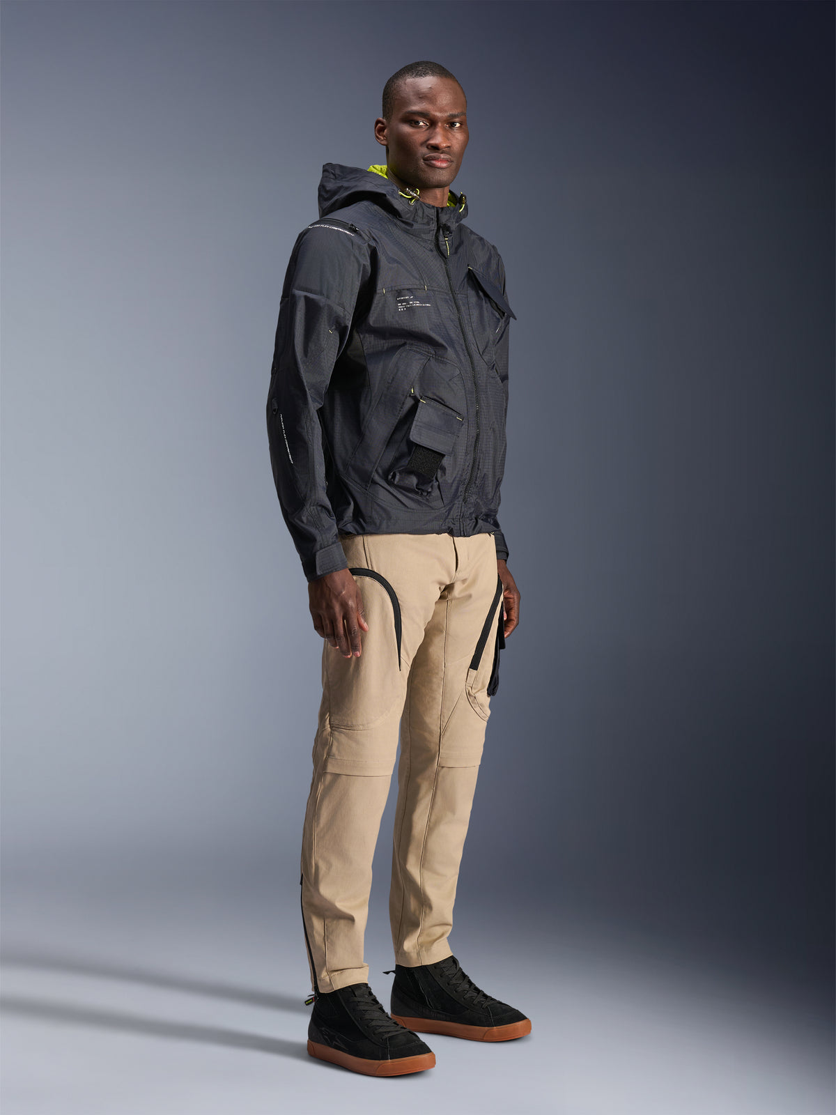 Pantalones Alpinestars Acteon, pantalones de moto urbanos, color Arena, lucidos por un modelo con una chaqueta técnica oscura y zapatillas negras, con un corte ajustado, detalles de cremalleras negras y bolsillos cargo, diseño contemporáneo inspirado en el motociclismo