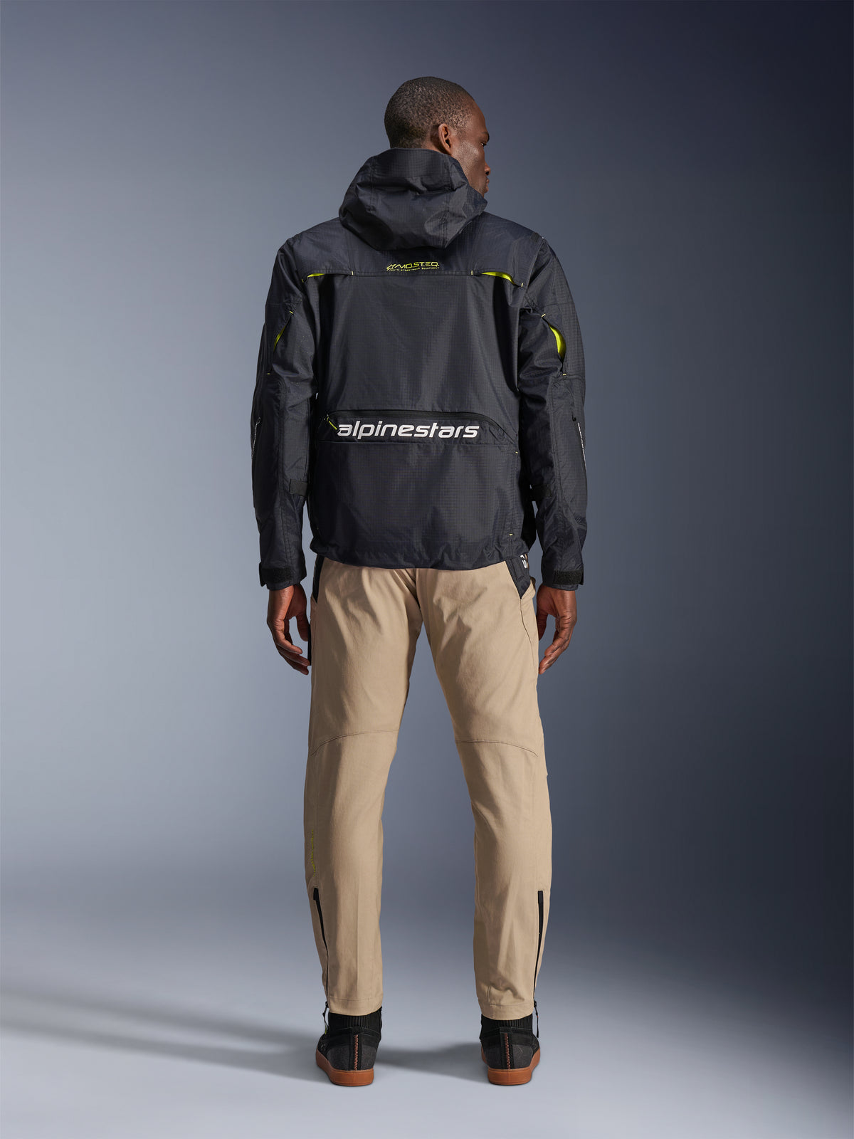 Pantalones Alpinestars Acteon, pantalones urbanos para moto, color arena, vistos desde atrás en un modelo que viste una chaqueta técnica negra con capucha y botas oscuras, con pierna cónica con cremalleras en los tobillos y una silueta limpia y moderna.