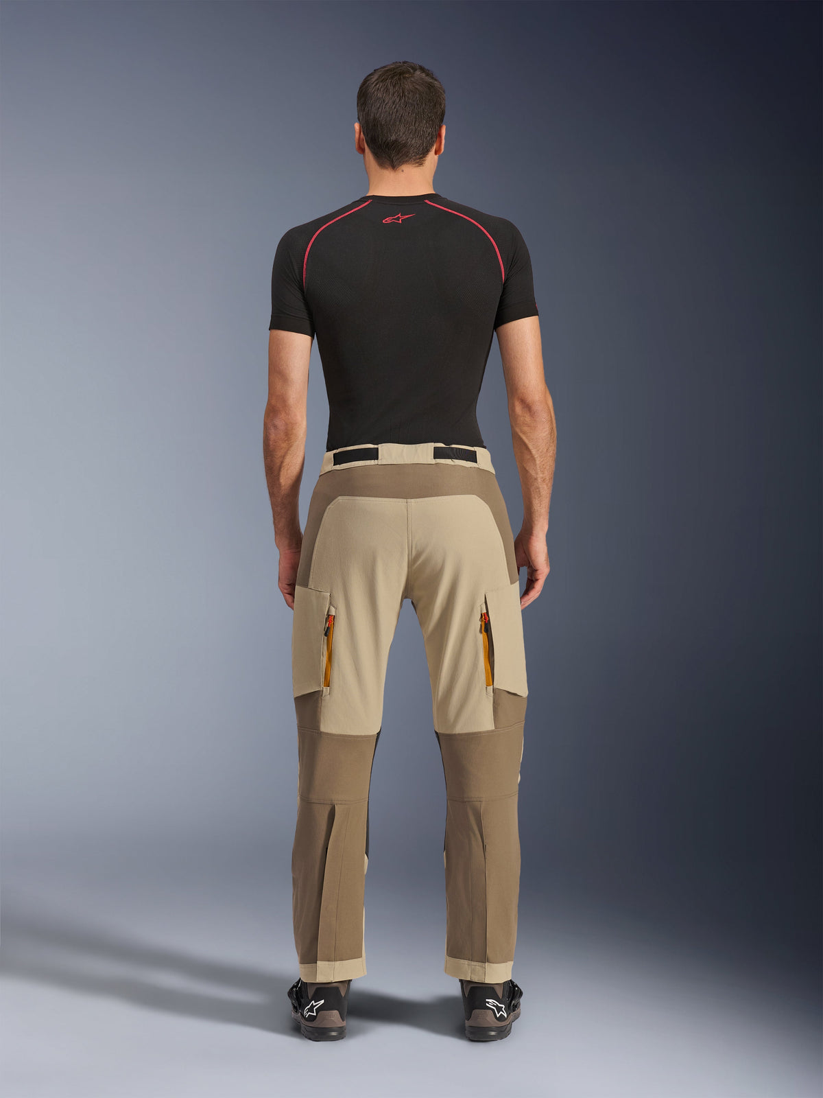 Pantalones Alpinestars Maxdura Dual en Walnut Aluminum, vista trasera que muestra un diseño bitono en marrón y tostado, pantalones para conducción de aventura con bolsillos de carga con cremallera y asiento reforzado, usados con una camiseta de compresión negra y botas de motociclismo.