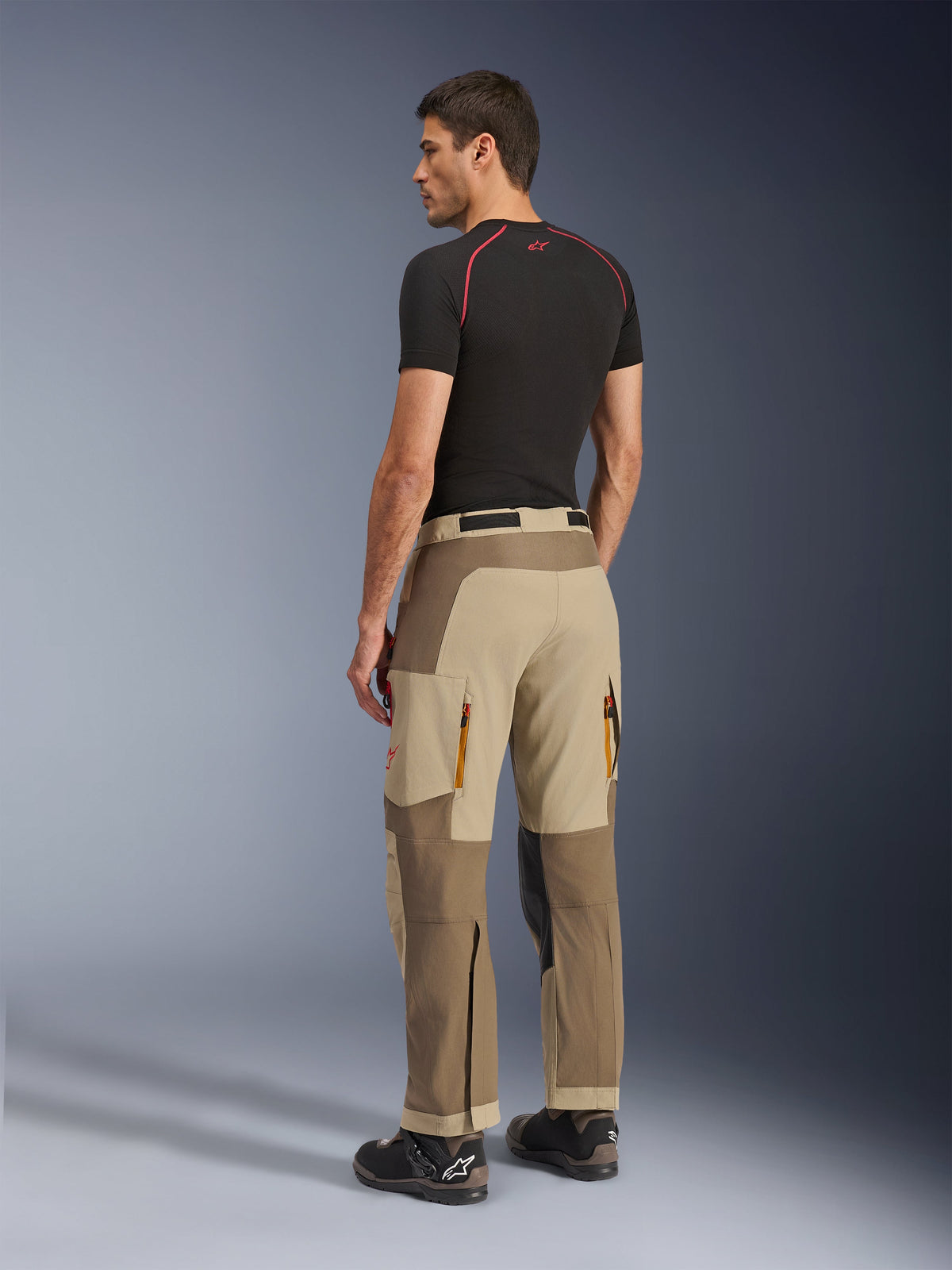 Pantalones Alpinestars Maxdura Dual, pantalones de conducción Adventure y Dual-Sport, Walnut Aluminum, vista trasera usados por un modelo con una capa base negra y botas de moto, con construcción de paneles múltiples, ventilaciones traseras con cremallera y refuerzos superpuestos.