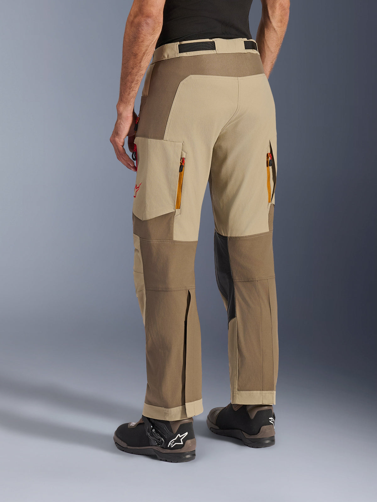 Pantalones Alpinestars Maxdura Dual en Walnut Aluminum, vista lateral trasera, pantalones de motociclismo de aventura con paneles en color canela y marrón, ventilaciones traseras con cremallera, grandes bolsillos de carga y diseño sobre la bota, combinados con botas negras y marrones