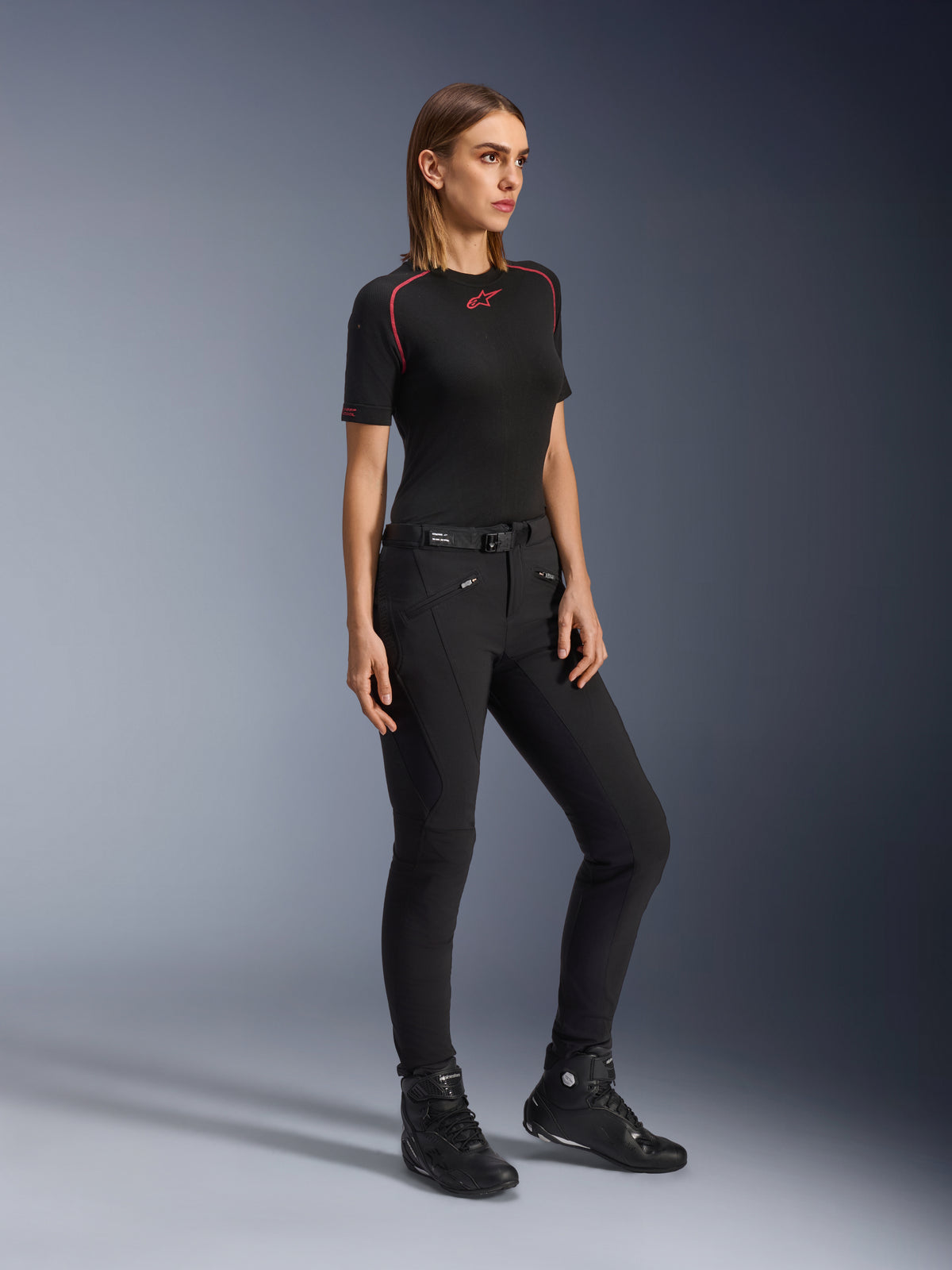Alpinestars Alamosa Women Pants, slim-fit motorcycle riding pants in black, worn by a model with a black base layer and riding shoes, featuring zippered front pockets and a magnetic closure waist belt, urban streetwear style.
