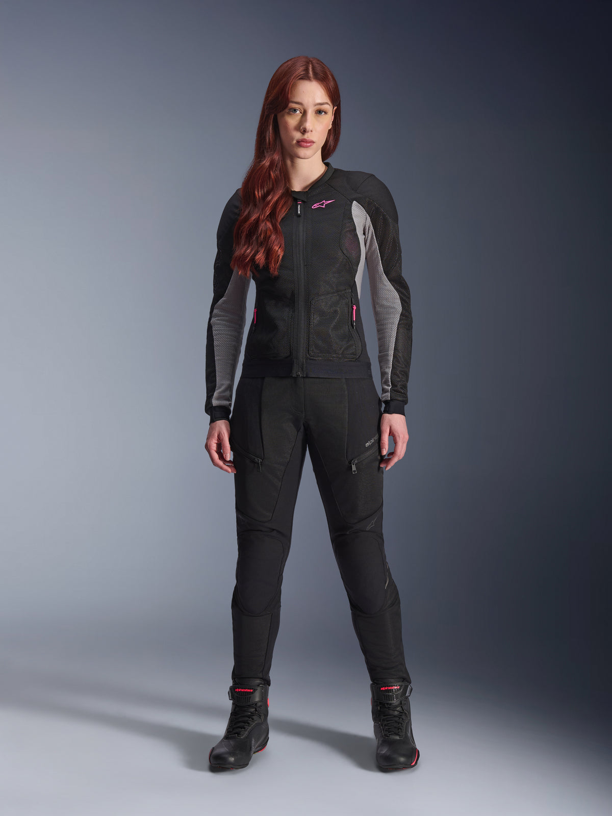 Alpinestars Stella Troop-Air Jacket and Pants, equipamiento de motociclismo para mujer, negro con detalles en gris ceniza y rosa flúor, usado por una modelo pelirroja, con ventilación de malla y diseño protector, pose de pie en estudio.