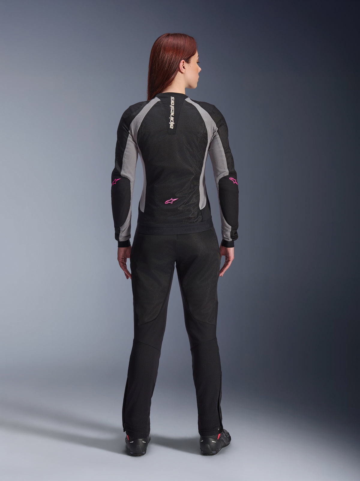 Chaqueta Stella Troop-Air para mujer y pantalones a juego vistos desde atrás, negro, gris ceniza y rosa flúor, equipamiento ligero de malla para motocicleta con paneles grises y detalles en rosa flúor, con la marca Alpinestars en vertical y logotipos de estrella.