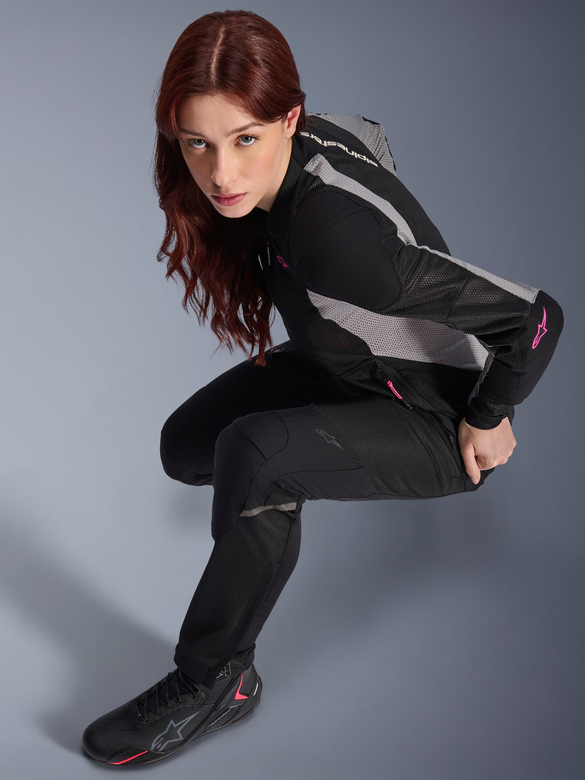 Mujer con pantalones Alpinestars Stella Troop-Air negros, pantalones textiles ligeros y ventilados, combinados con una chaqueta de malla negra y gris con detalles rosas y botas de moto negras, retrato de estudio, equipamiento para conducción urbana en moto
