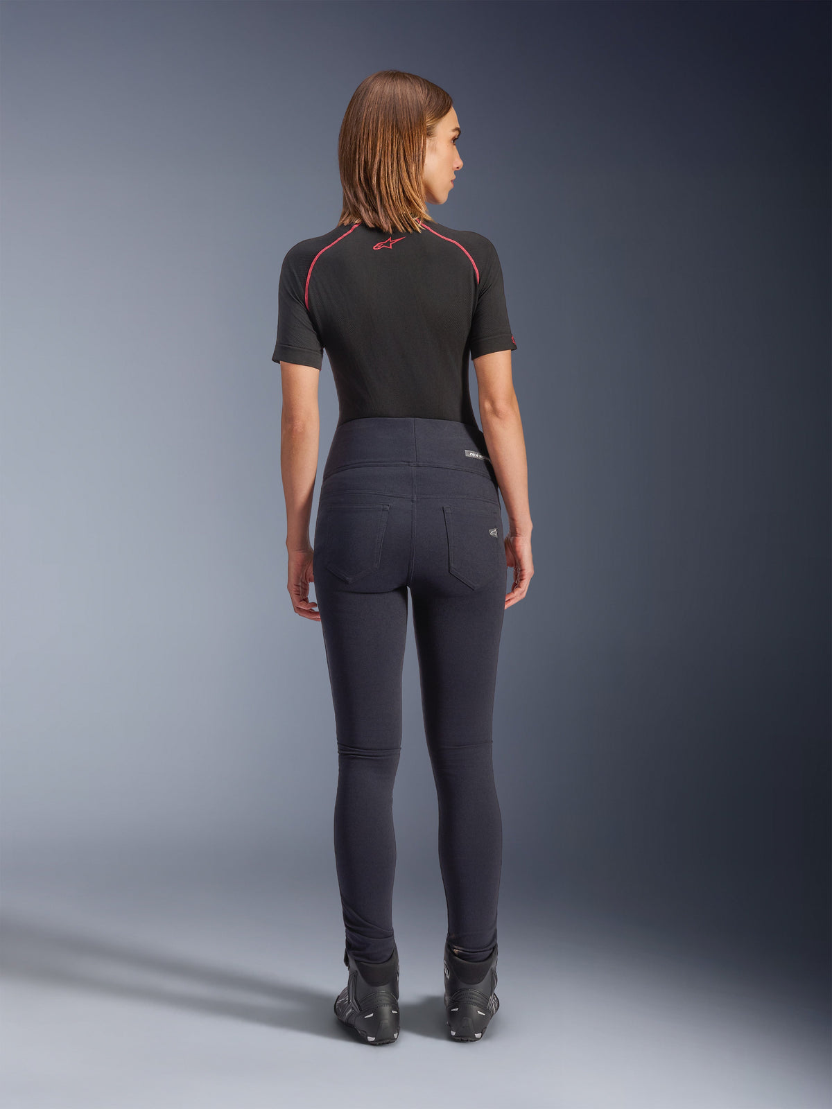 Leggings Alpinestars Stella Flex-AST en negro, pantalones de moto de talle alto para mujer, vista trasera que muestra el ajuste ceñido y los bolsillos traseros, combinados con una camiseta negra de manga corta y botas de conducción, equipamiento de conducción urbana