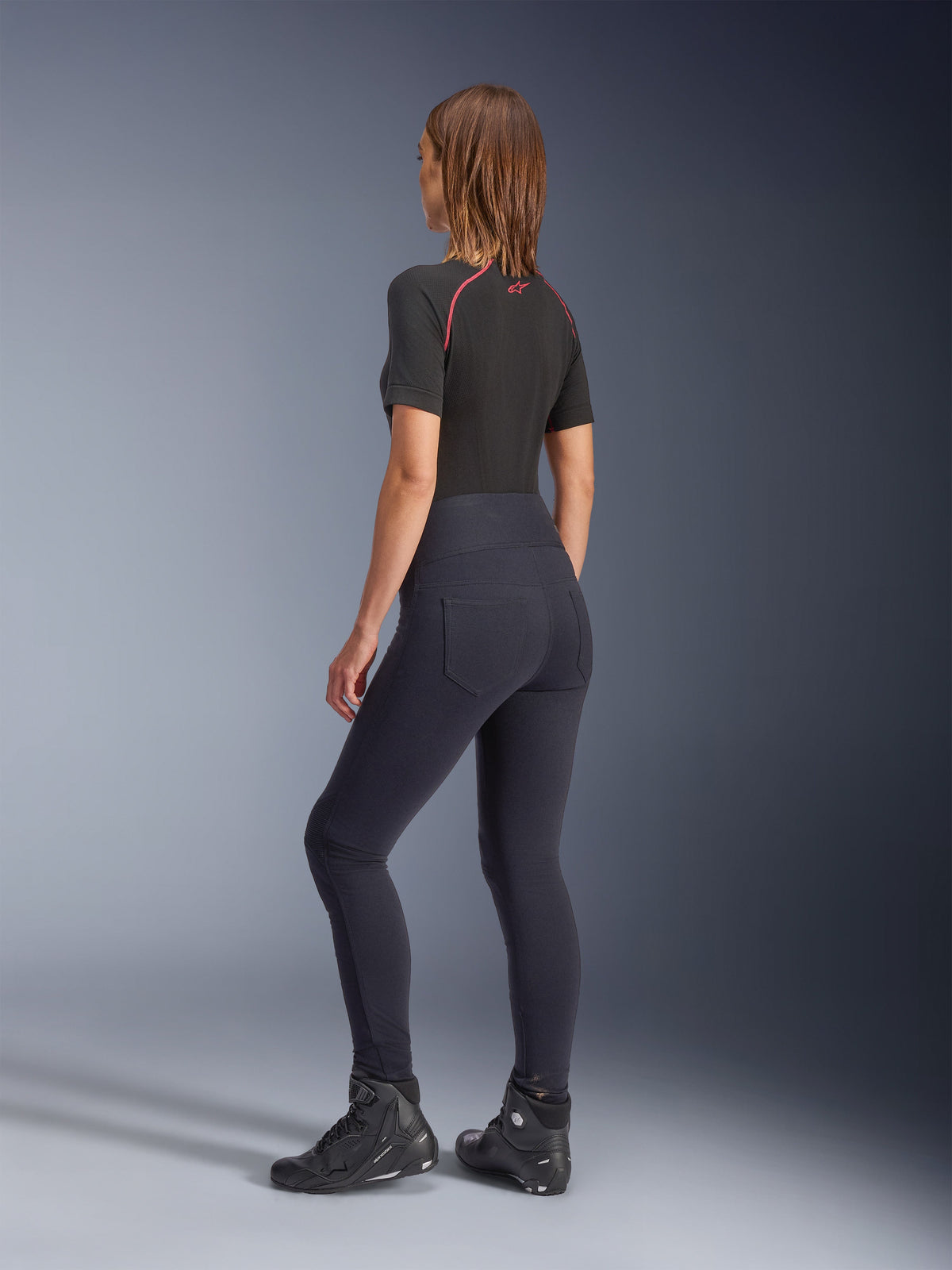 Leggings Alpinestars Stella Flex-Ast para mujer, negros, vista trasera que muestra el ajuste de cintura alta y los bolsillos traseros, combinados con una prenda base de manga corta negra con detalles rojos y calzado de moto negro, equipamiento contemporáneo para moto de estilo urbano
