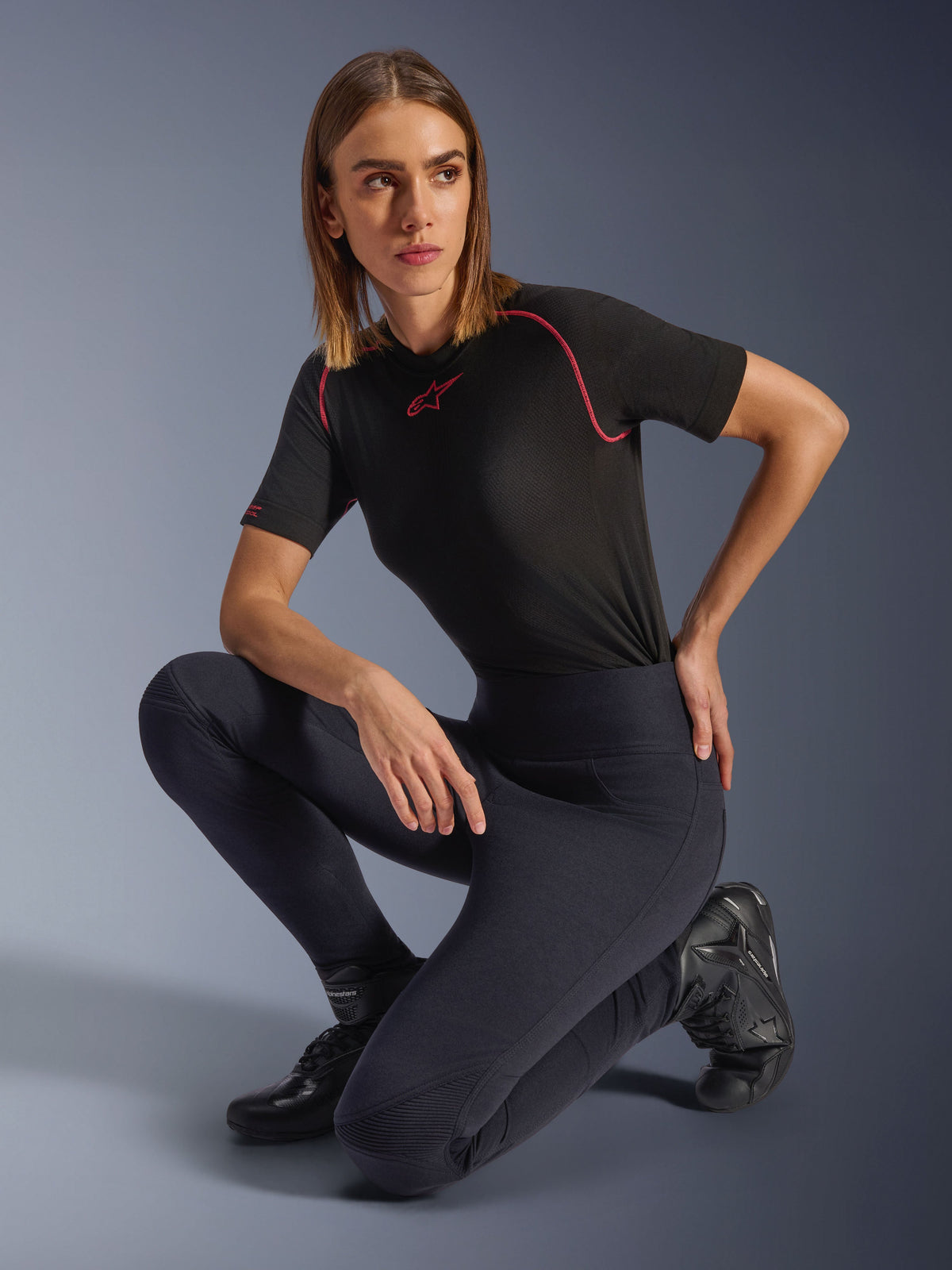 Mujer vistiendo leggings Stella Flex-AST de Alpinestars, leggings de moto negros ajustados de talle alto con paneles de acordeón en las rodillas, combinados con una camiseta técnica de manga corta negra con costuras rosas y botas de conducción.