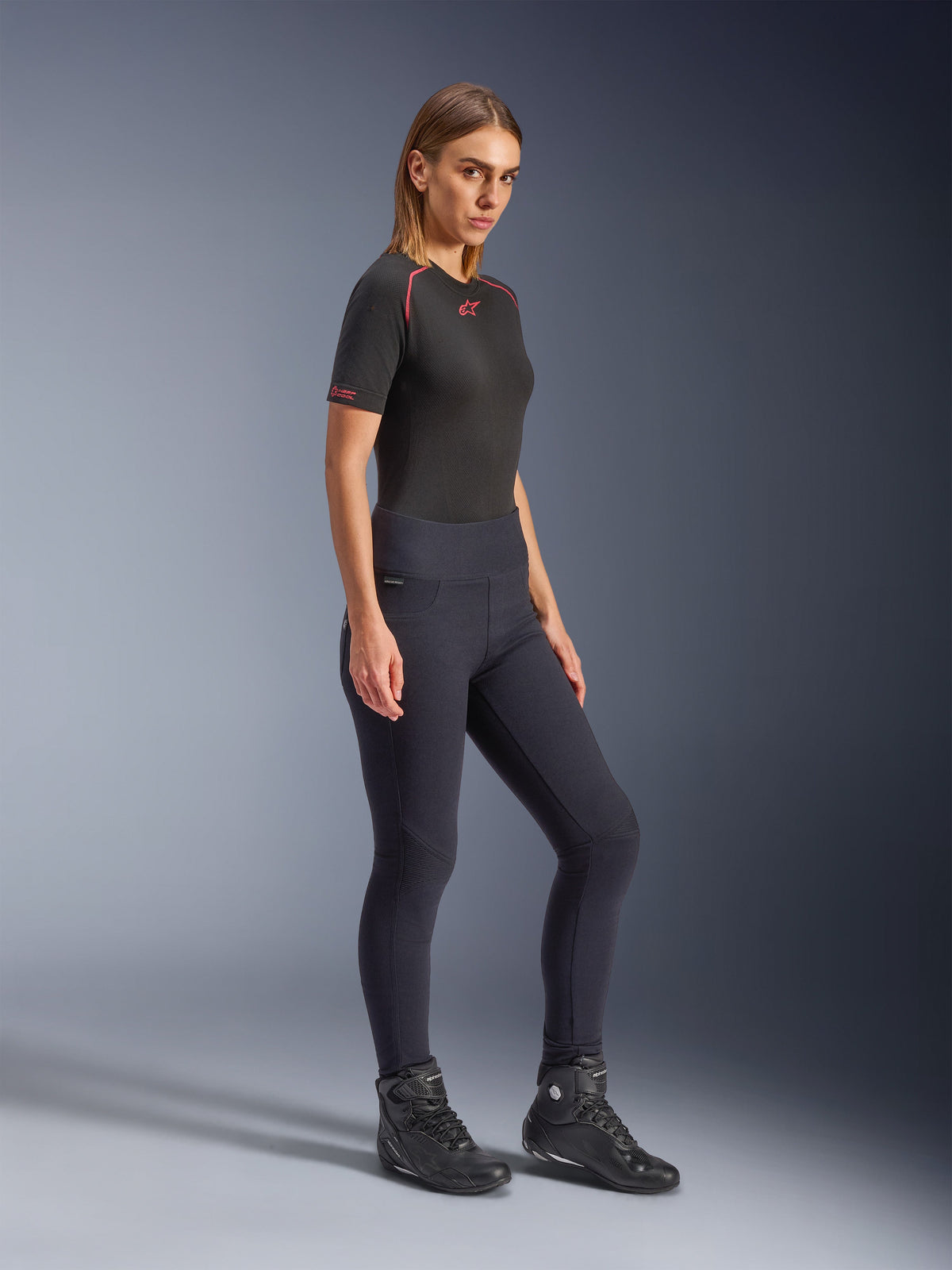 Alpinestars Stella Flex-Ast Leggings, leggings de moto para mujer en color negro, diseño de talle alto y ajuste ceñido, combinados con una camiseta negra con detalles rojos y botas de moto negras, toma de cuerpo completo en estudio