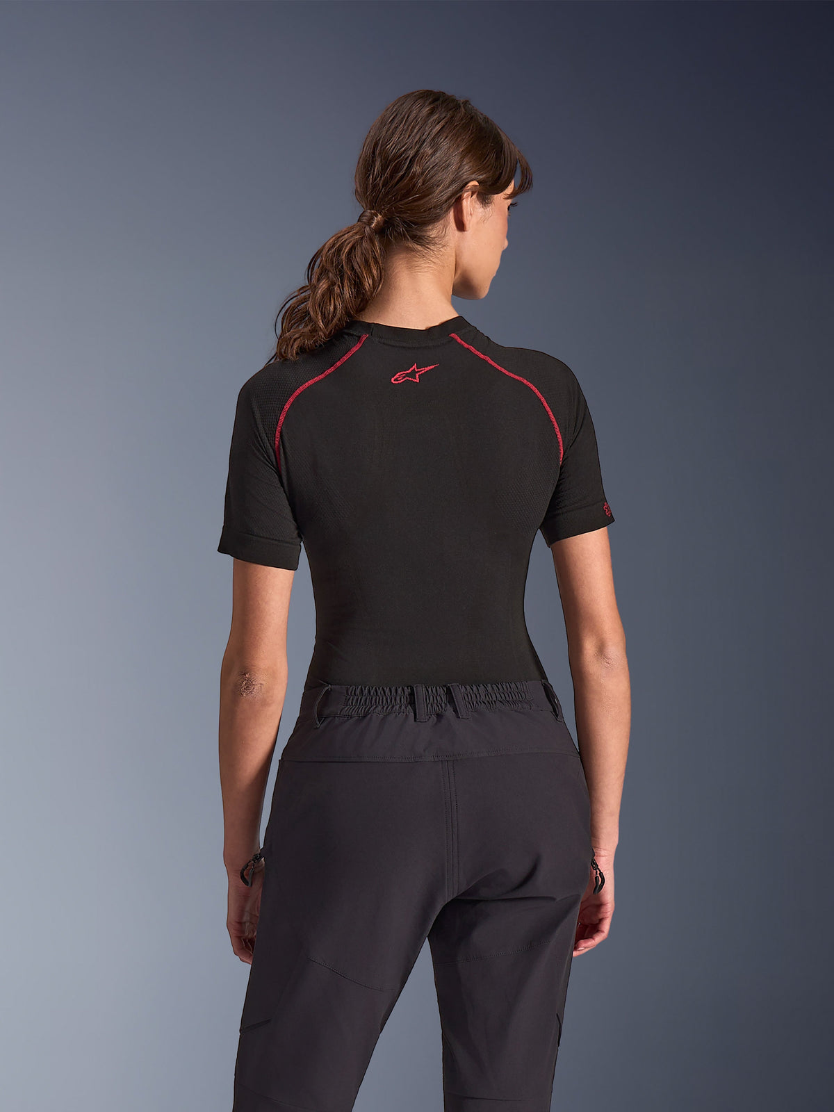 Woman wearing Alpinestars Stella Flex-AST Explorer Pants, Black adventure motorcycle pants, back view, paired with a black short-sleeved top with red stitching, highlighting the breathable textile construction and elastic waistband.